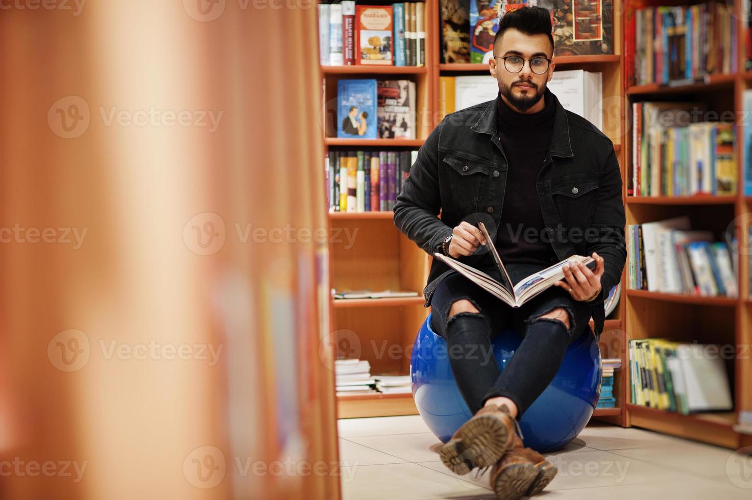Tall smart arab student man, wear on black jeans jacket and eyeglasses, sitting at library and read book at hands. photo