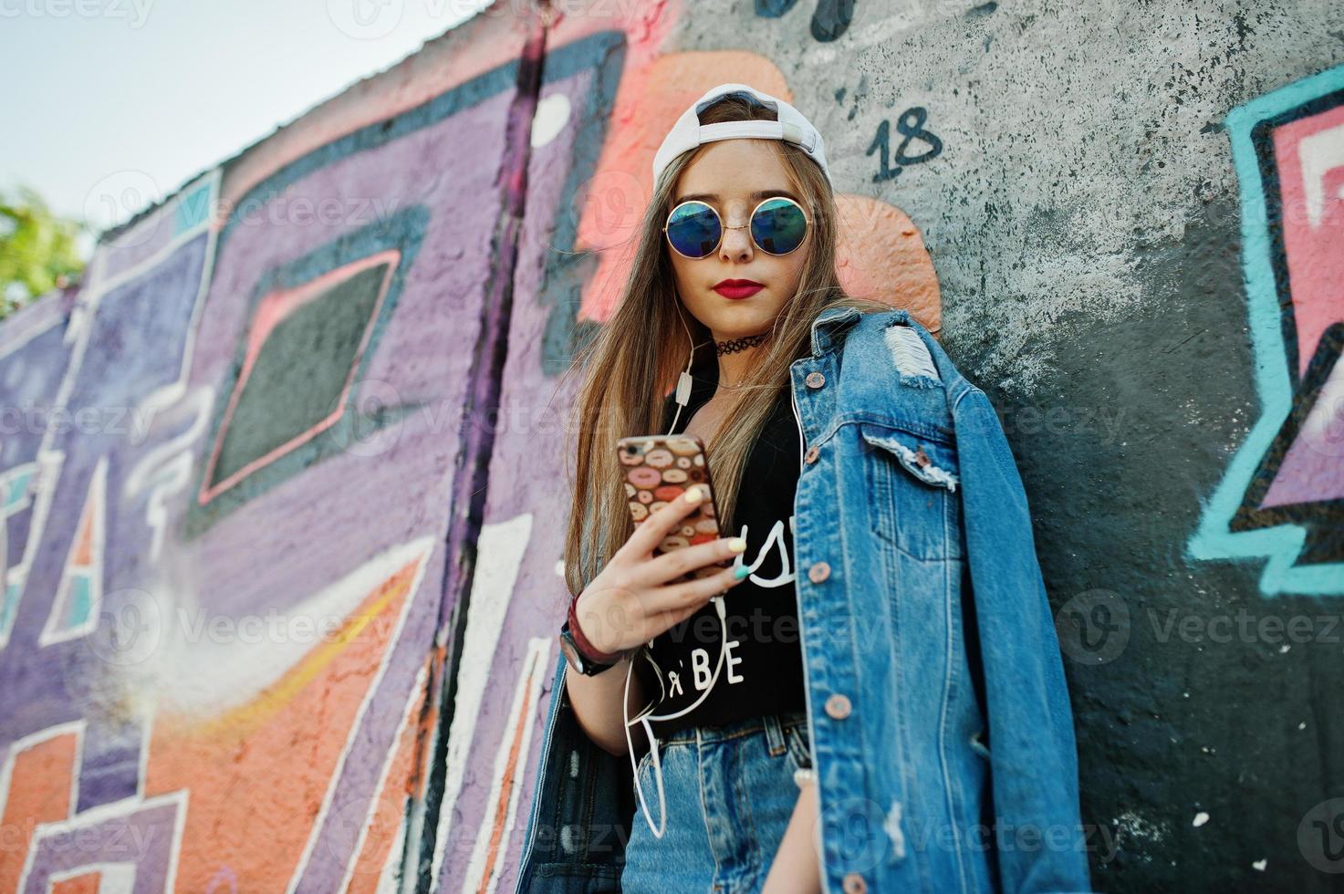 Stylish casual hipster girl in cap, sunglasses and jeans wear, listening music from headphones of mobile phone against large graffiti wall. photo