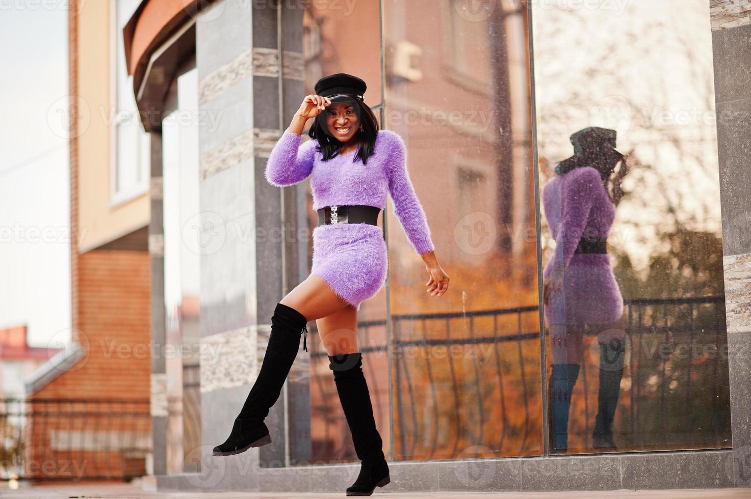 African american woman at violet dress and cap posed outdoor against modern building. photo