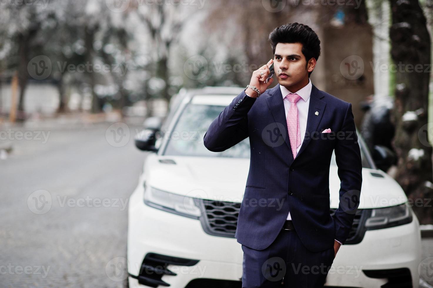Elegant indian macho man model on suit and pink tie posed against white business car and speaking on phone. photo