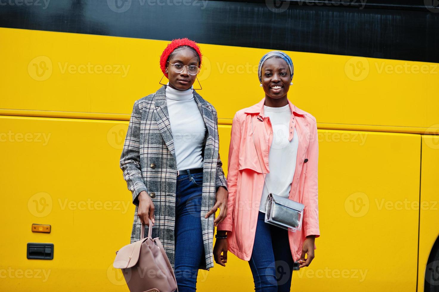 Two young modern fashionable, attractive, tall and slim african muslim womans in hijab or turban head scarf and coat posed against yellow bus. photo