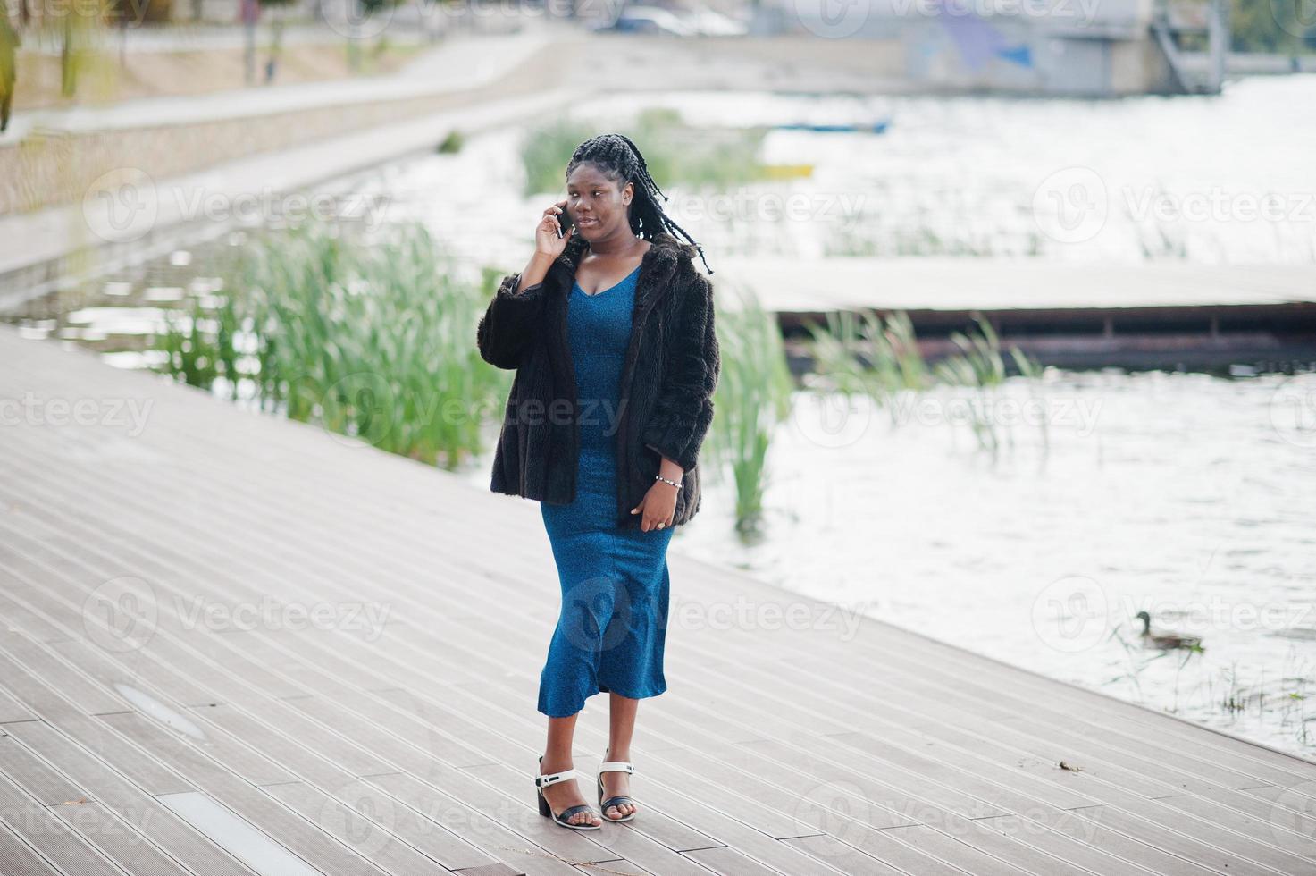 African american dark skinned plus size model posed in a blue shiny dress and black fur coat against lake and speaking by mobile phone. photo