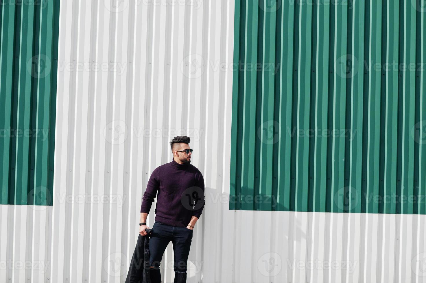 Fashion rich beard Arab man wear on turtle neck and sunglasses posed against green and white steel wall with jacket on hand. Stylish, succesful and fashionable arabian model guy. photo