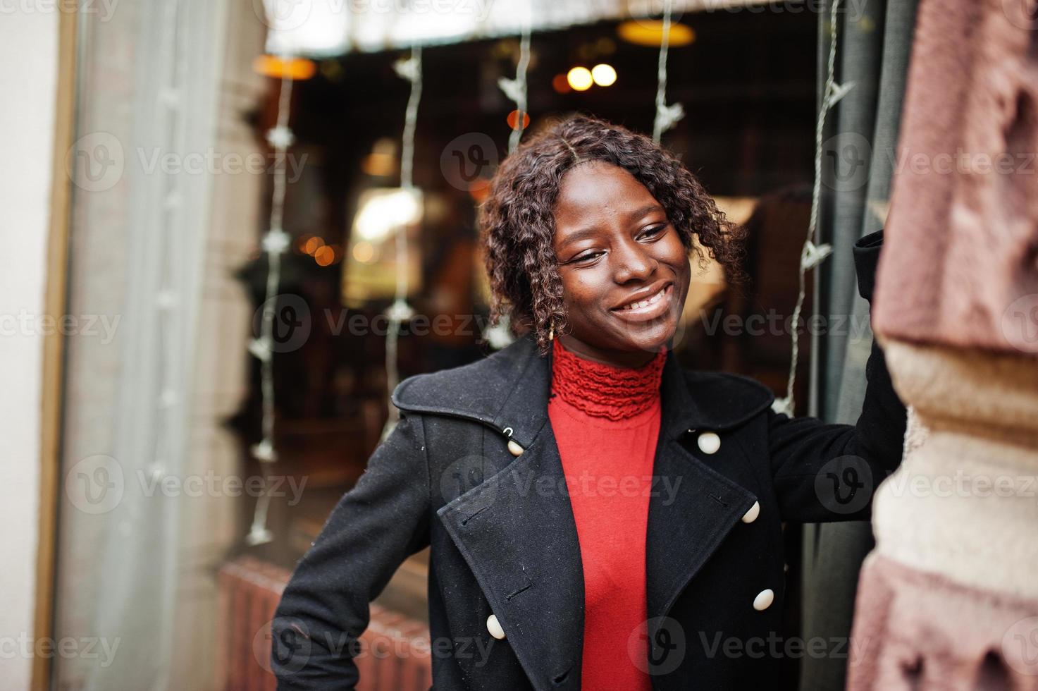 retrato de una mujer africana de pelo rizado con un abrigo negro de moda y un cuello alto rojo posando al aire libre. foto