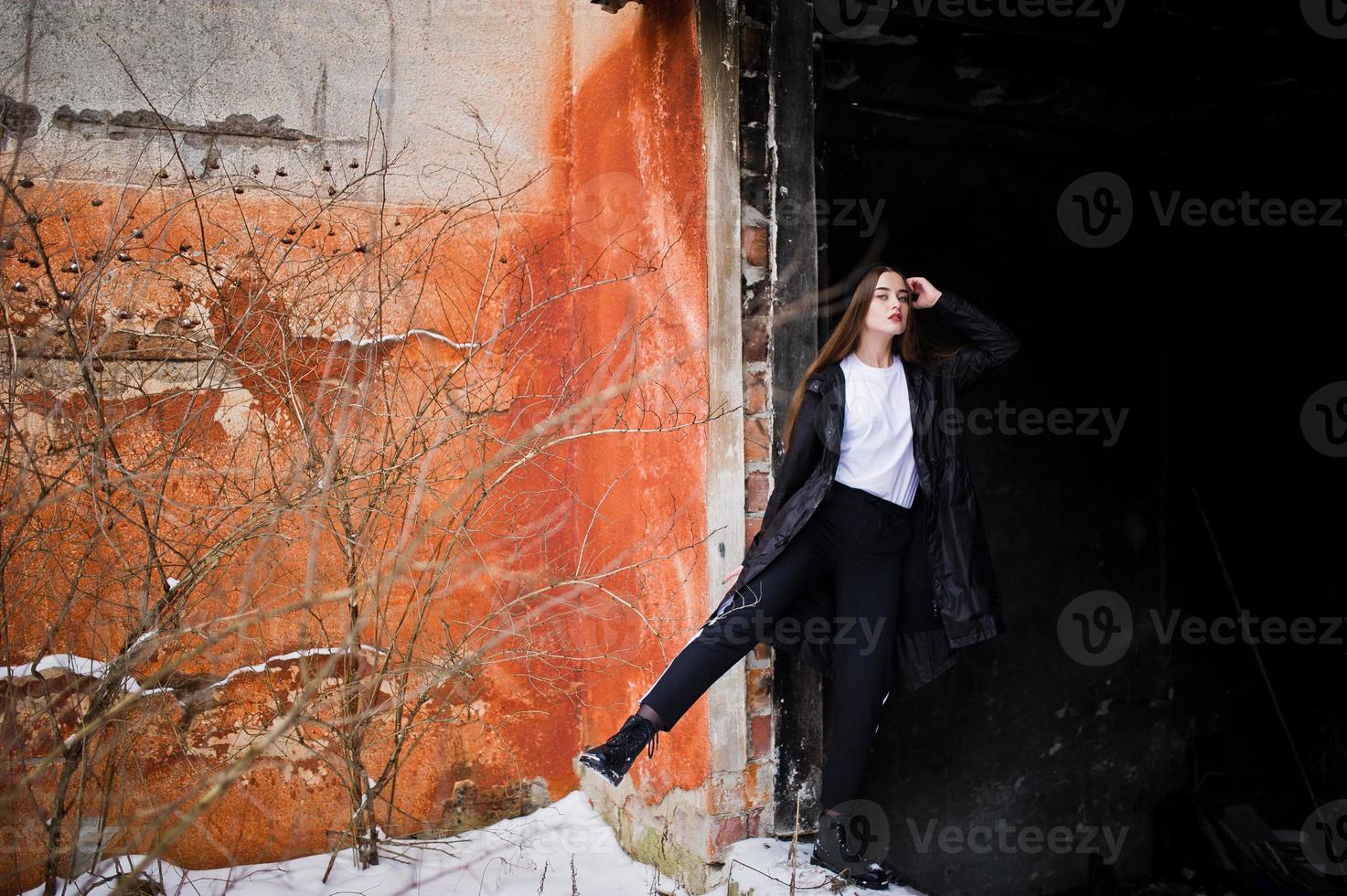 modelo morena de piernas largas de moda en un largo manto negro posado al aire libre en el día de invierno contra la vieja pared del grunge. foto