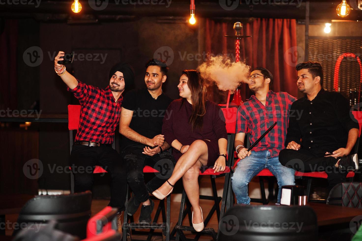 Group of indian friends sitting at lounge bar, having fun and rest, smoking hookah and looking on mobile phones to make selfie. photo