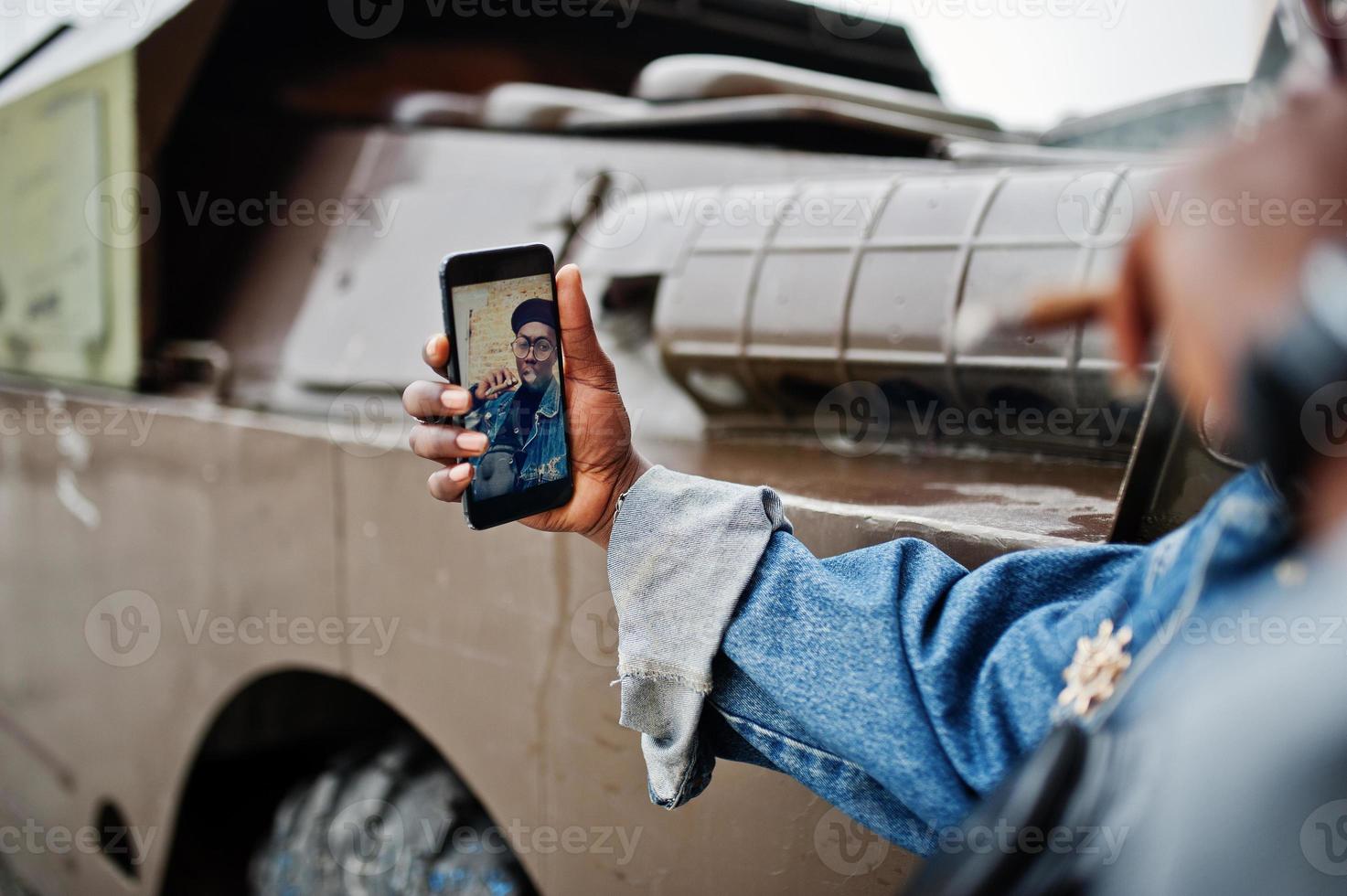 African american man in jeans jacket, beret and eyeglasses, smoking cigar and posed against btr military armored vehicle, making selfie on phone. Focus on screen of cellphone. photo