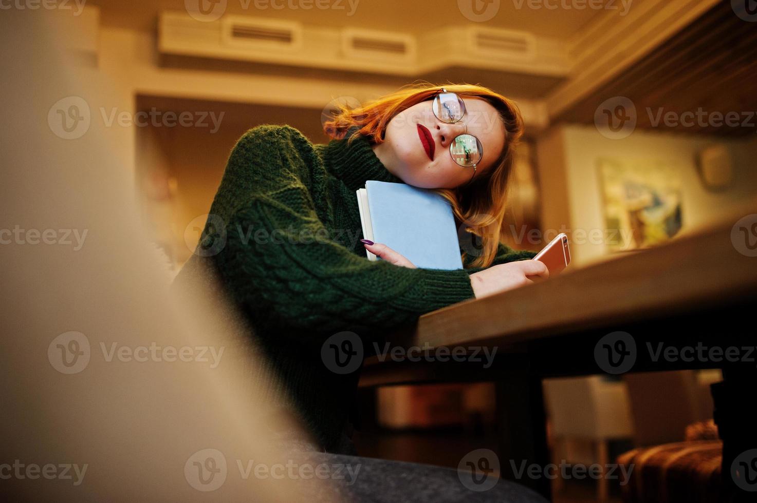 Dreaming about something beautiful. Cheerful young beautiful redhaired woman in glasses, green warm wool sweater, using her phone and notebook, while sitting at her working place on cafe. photo