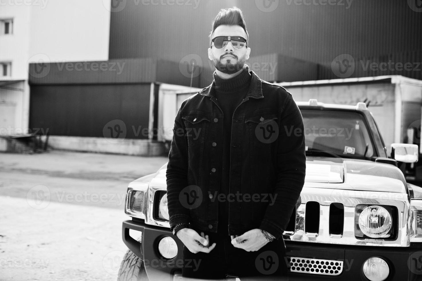 El hombre árabe de barba rica en moda usa una chaqueta negra de jeans y gafas de sol posadas contra un gran auto todoterreno negro. chico modelo árabe elegante, exitoso y de moda. foto