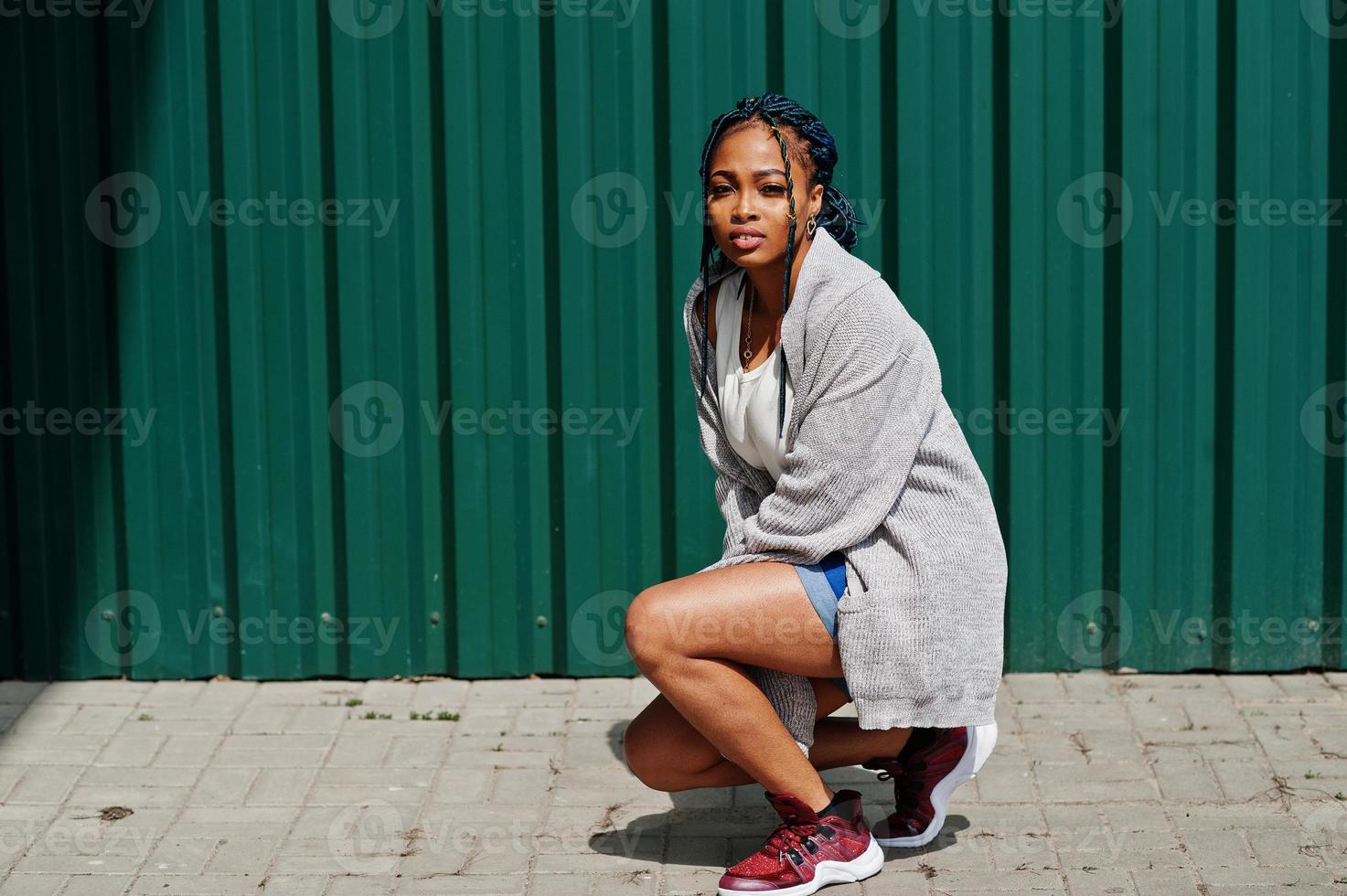 African woman with dreads hair, in jeans shorts crouching against green steel wall. photo