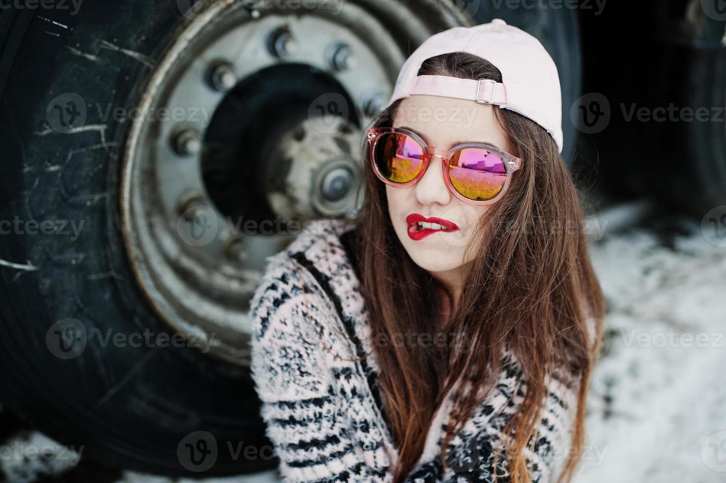 Brunette stylish casual girl in cap and sunglasses sitting against truck wheels. photo