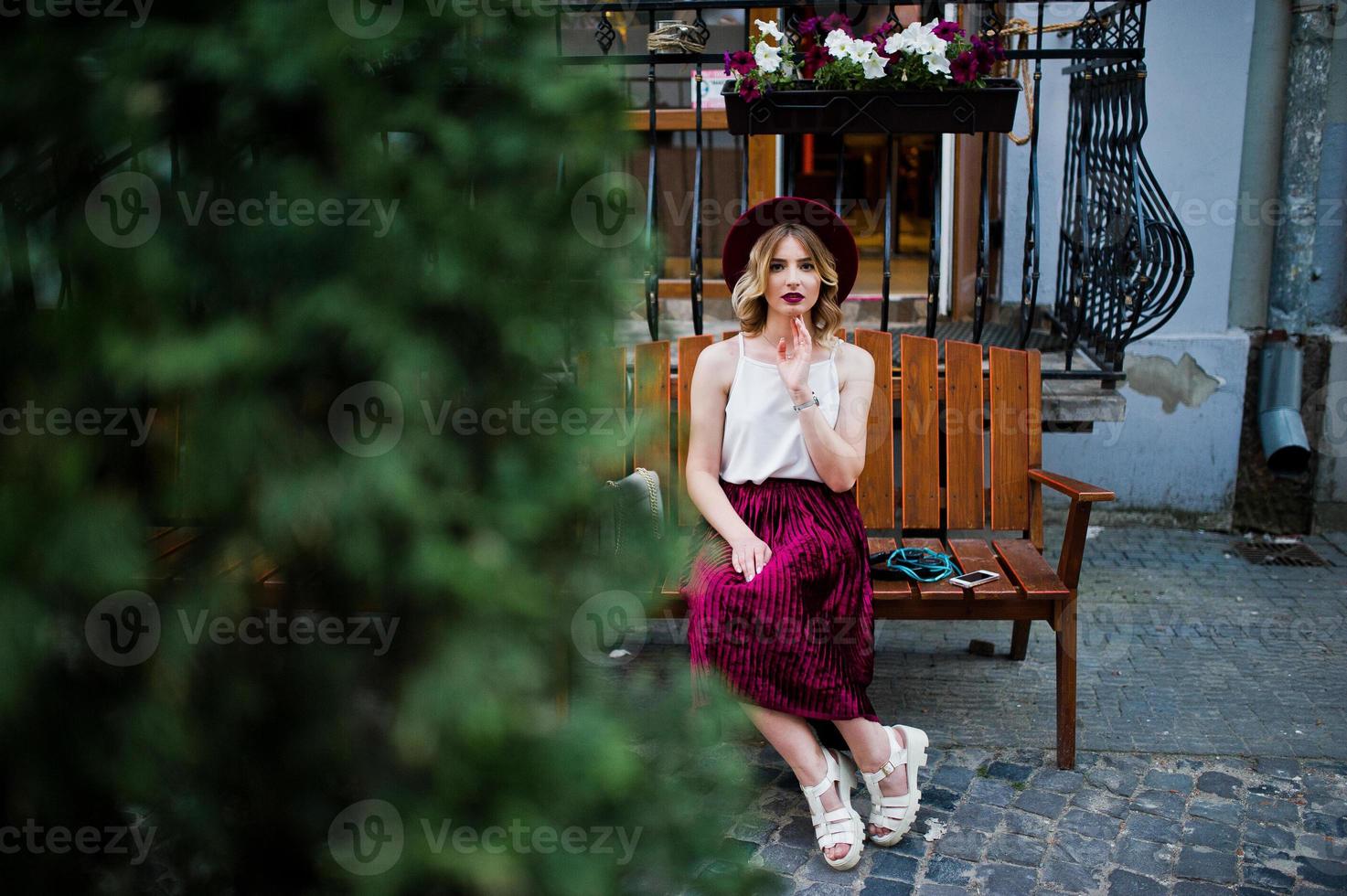 Fashionable and beautiful blonde model girl in stylish red velvet velour skirt, white blouse and hat, posed on bench at city street. photo