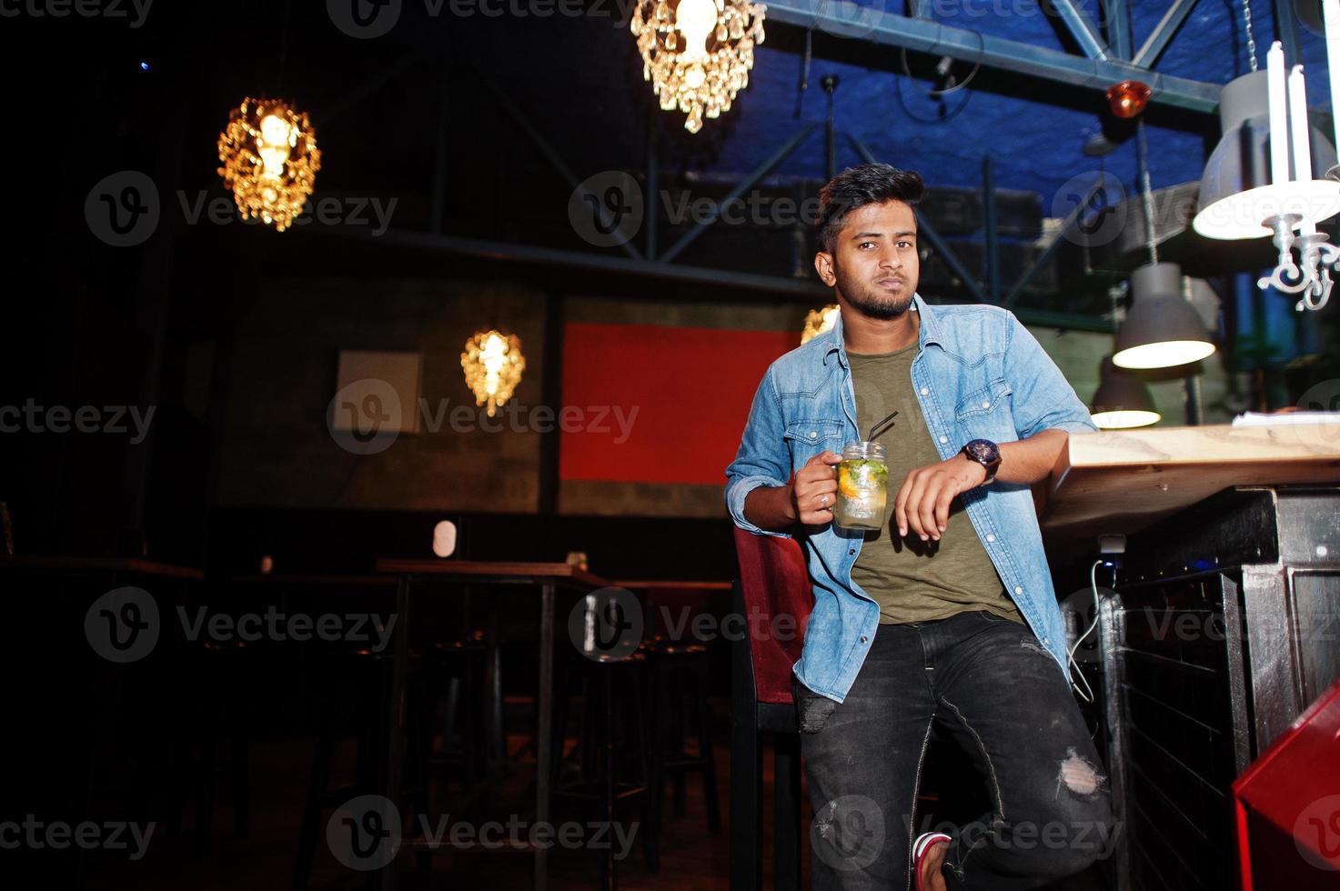 retrato de un apuesto y exitoso surasiático barbudo, joven autónomo indio con camisa de jeans azul sentado en un club nocturno contra el mostrador del bar con un cóctel y descansando. foto