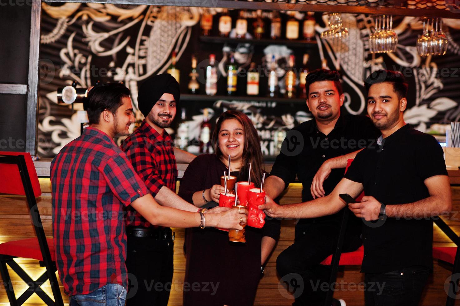 Group of indian friends having fun and rest at night club, drinking cocktails, clinking and cheers near bar counter. photo