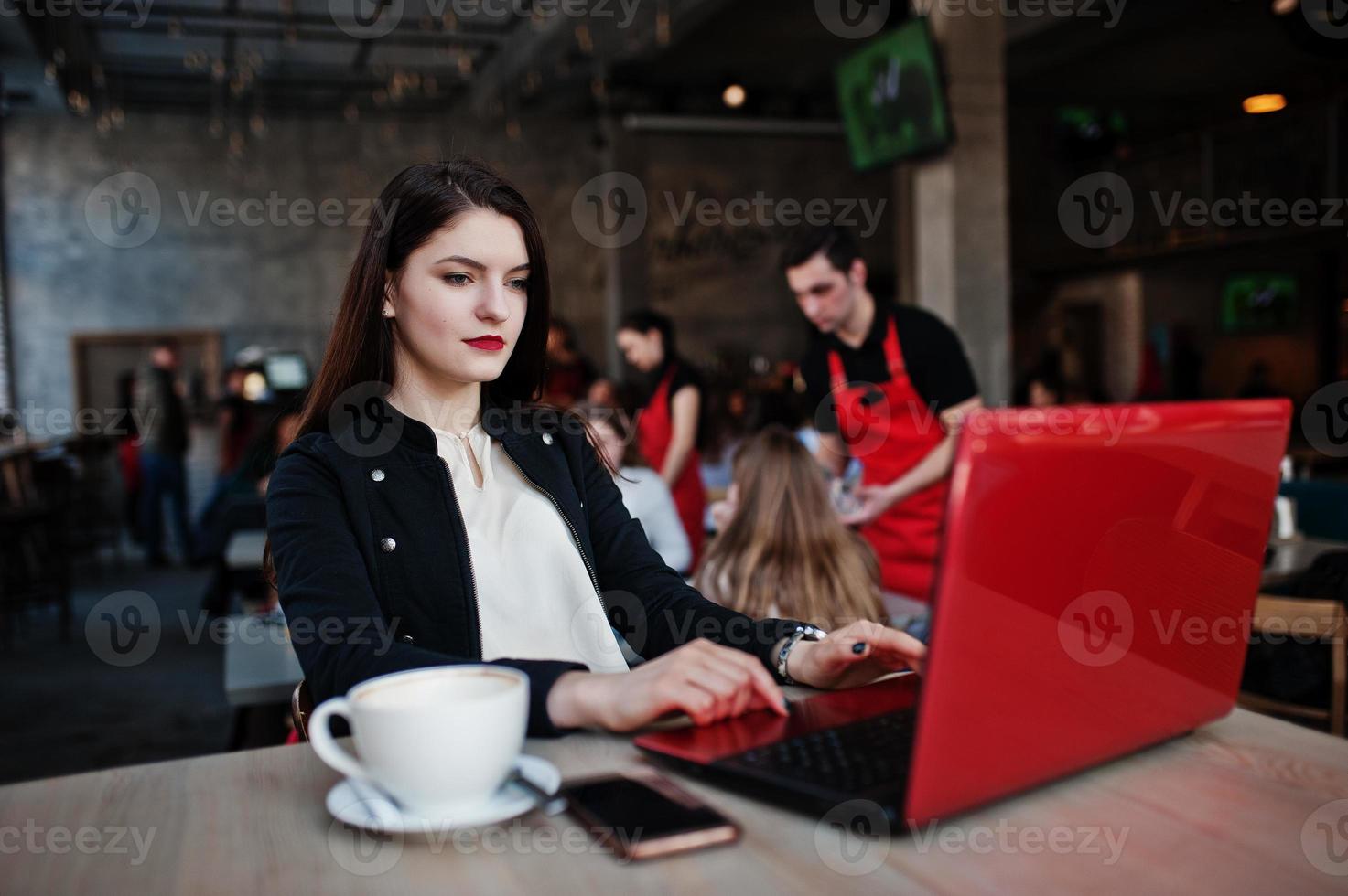 chica morena sentada en un café con una taza de capuchino, trabajando con una laptop roja. foto