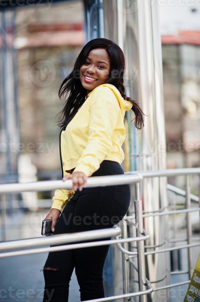 Hipster african american girl wearing yellow hoodie posing at street against office building with blue windows. photo