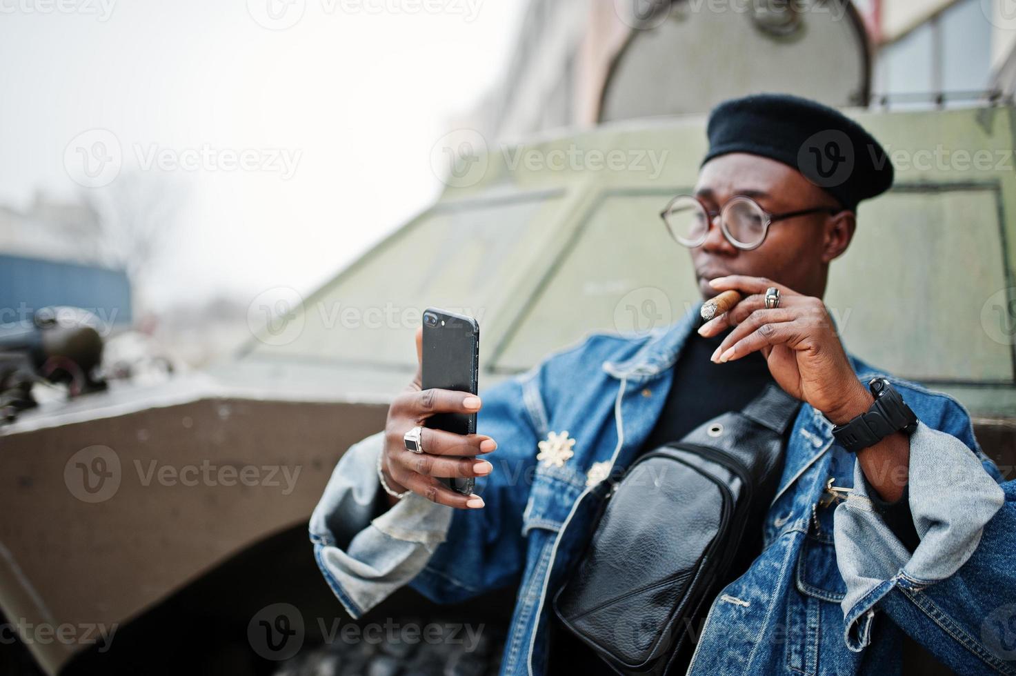 African american man in jeans jacket, beret and eyeglasses, smoking cigar and posed against btr military armored vehicle, making selfie on phone. photo