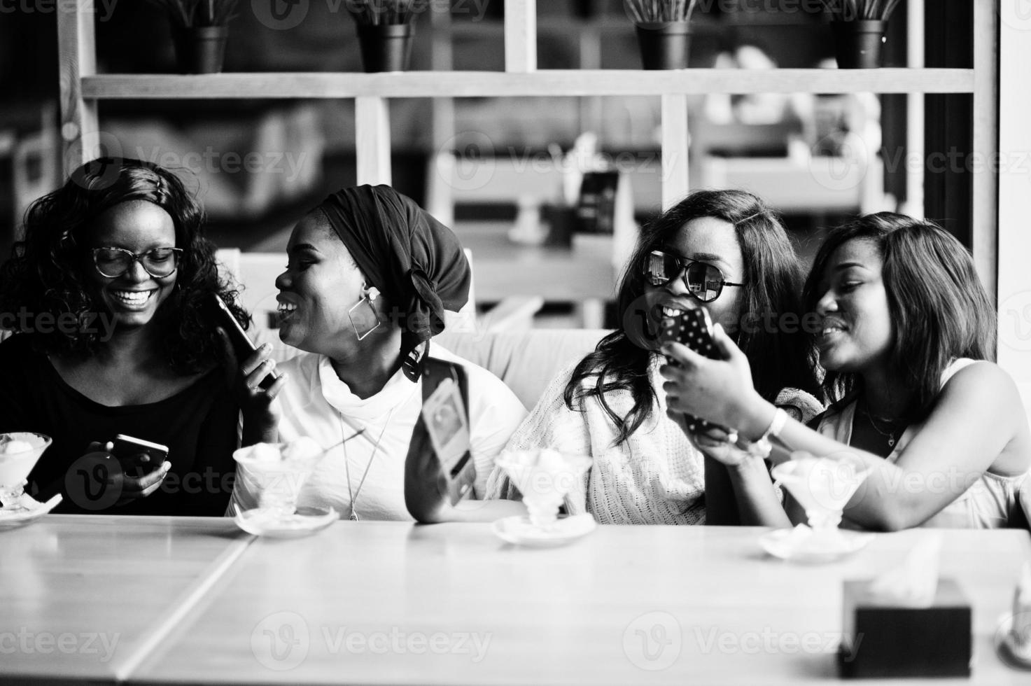 cuatro chicas afroamericanas sentadas en la mesa del café con postre helado y mirando fotos en los teléfonos móviles.