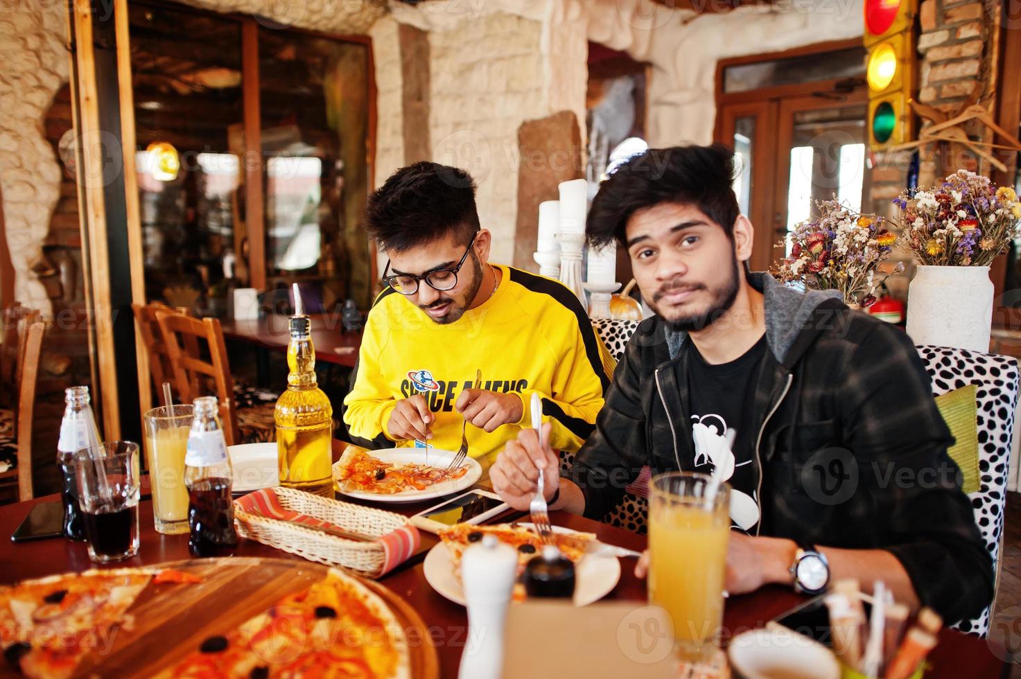 Asian friends guys eating pizza during party at pizzeria. Happy indian people having fun together, eating italian food and sitting on couch. photo