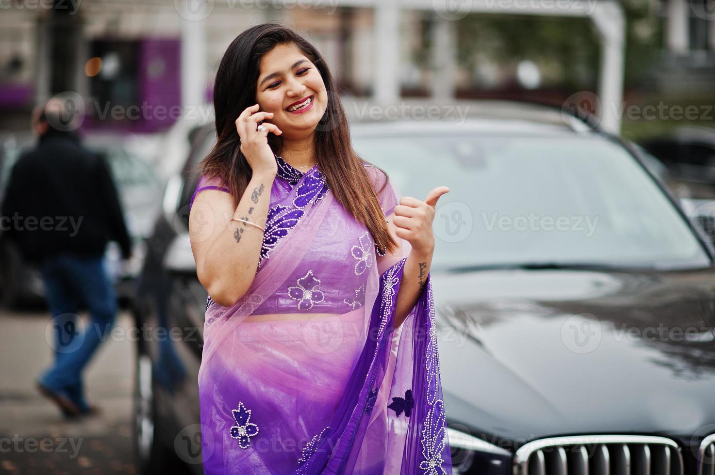 Indian hindu girl at traditional violet saree posed at street against black business suv car and speaking on mobile phone, shows thumb up. photo
