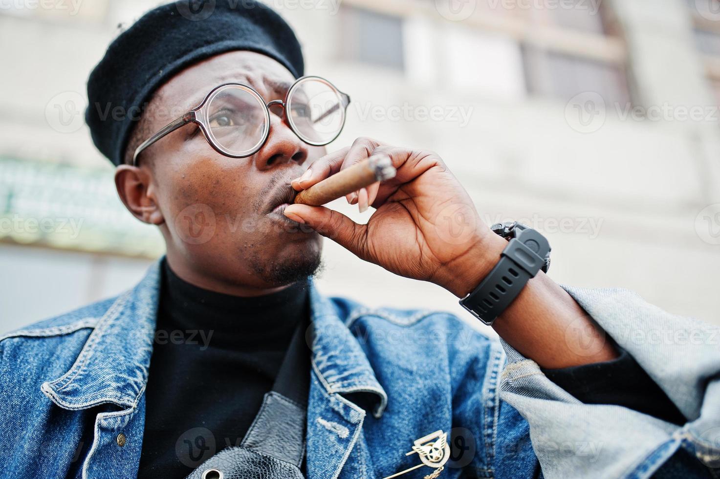 Close up portrait of african american man in jeans jacket, beret and eyeglasses, smoking cigar. photo