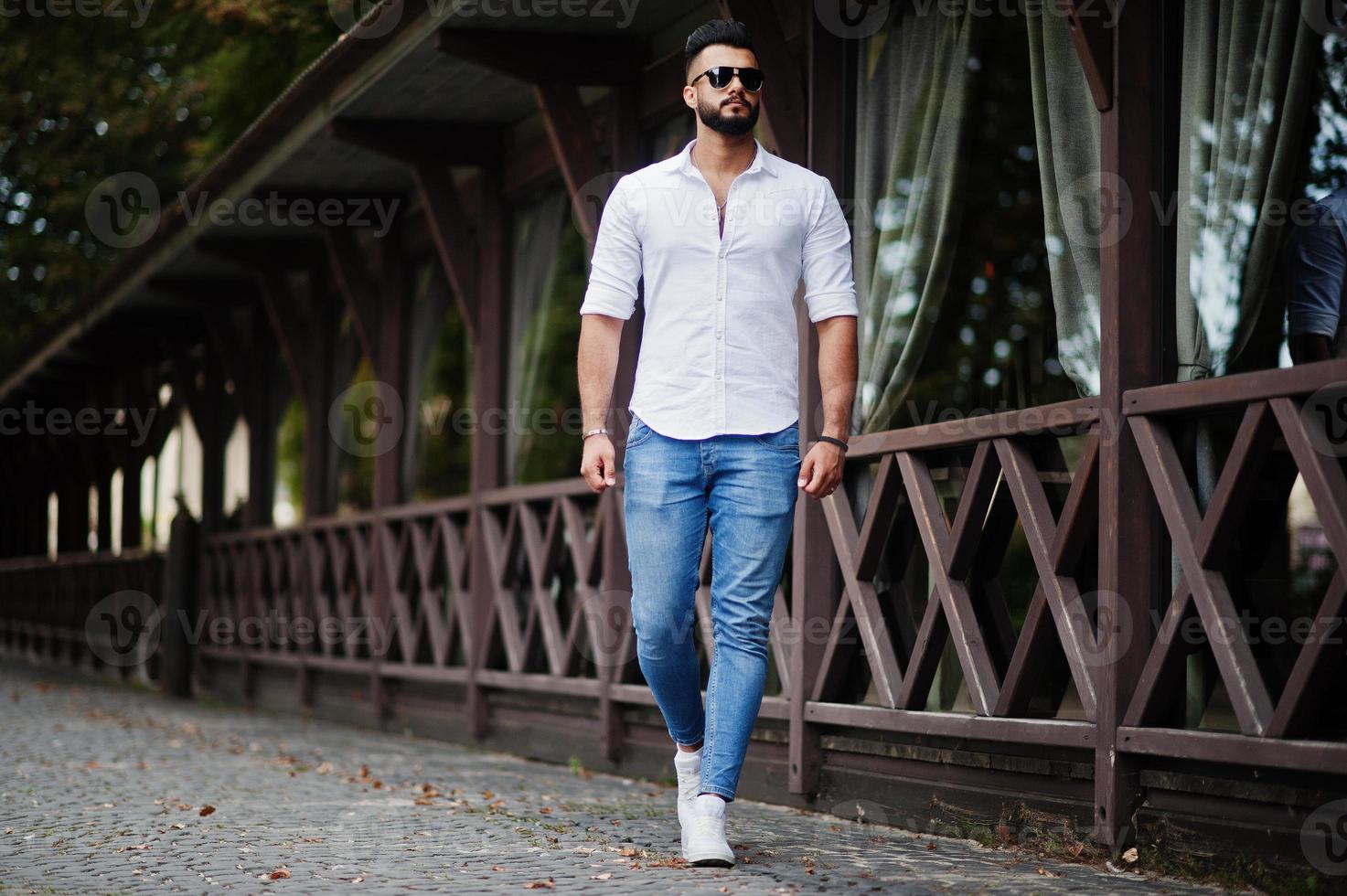 Stylish tall arabian man model in white shirt, jeans and sunglasses walking at street of city. Beard attractive arab guy. photo