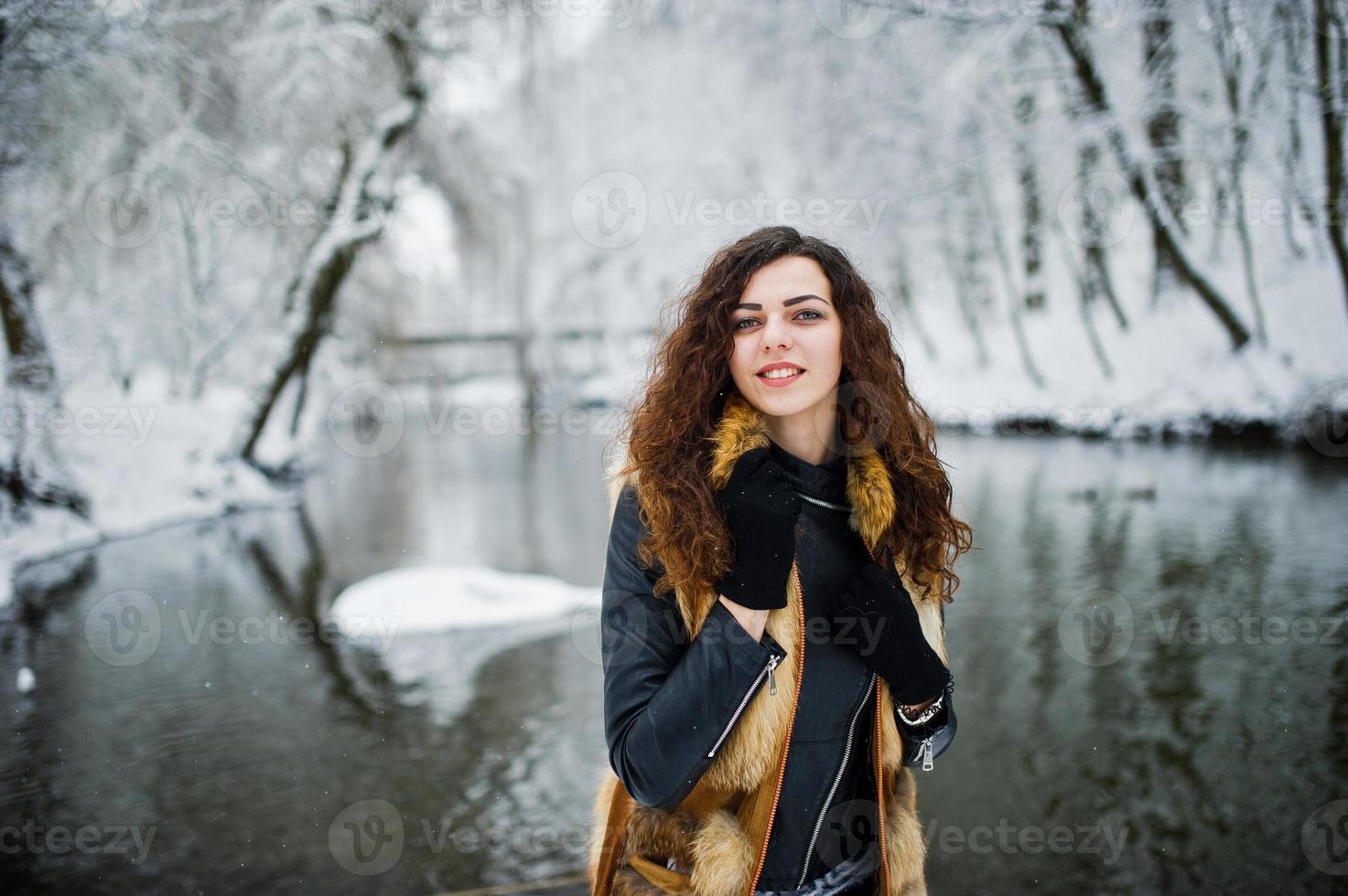 Elegance curly girl in fur coat at snowy forest park agasinst frozen river at winter. photo