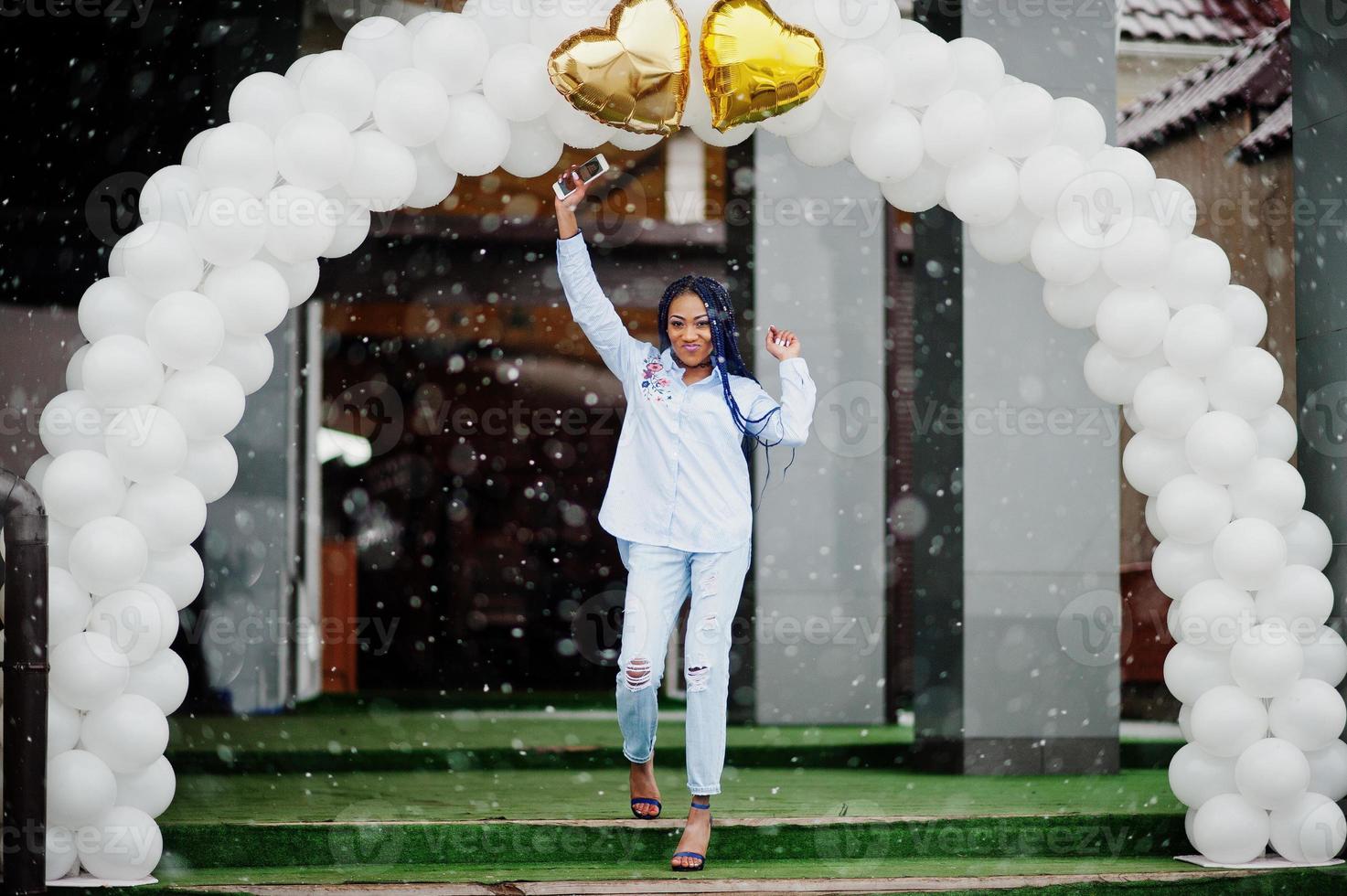 Stylish african american girl with dreads holding mobile phone at hand, outdoor against arch of balloons at snowy weather. photo