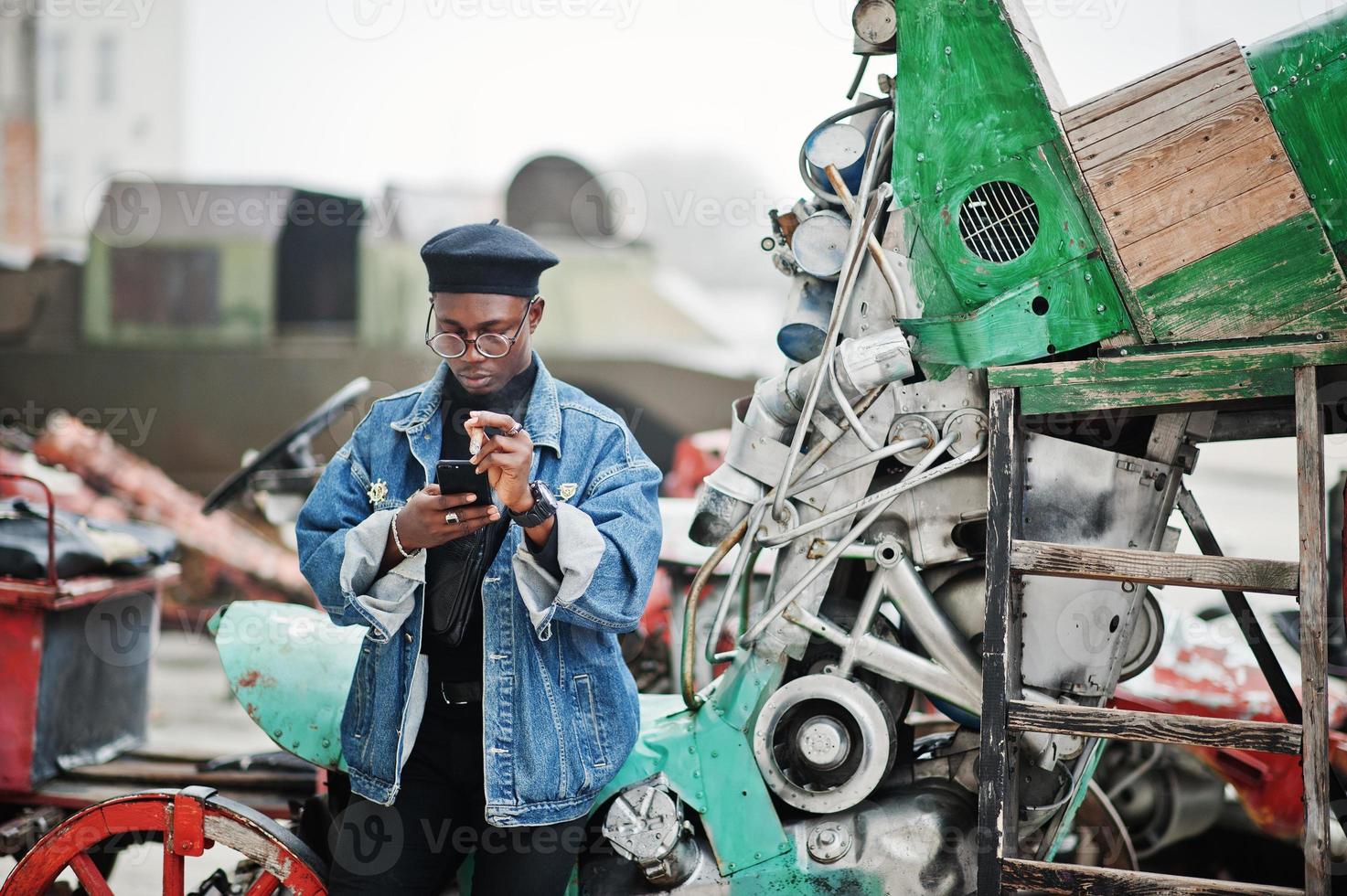 African american man in jeans jacket, beret and eyeglasses, smoking cigar and posed against old retro vehicle and looking at phone. photo