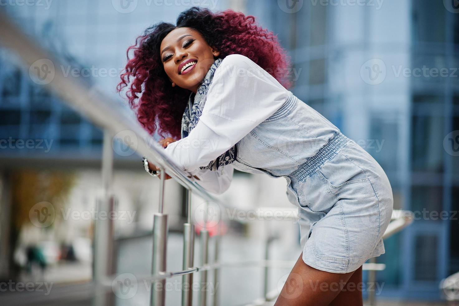 atractiva mujer afroamericana rizada vestida de jeans posada contra un moderno edificio de varios pisos. foto
