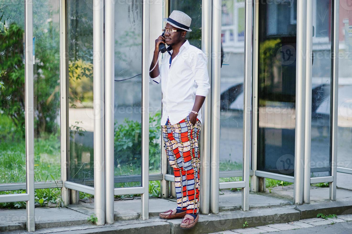 Stylish african american man in white shirt and colored pants with hat and glasses posed outdoor at telephone booth. Black fashionable model boy. photo
