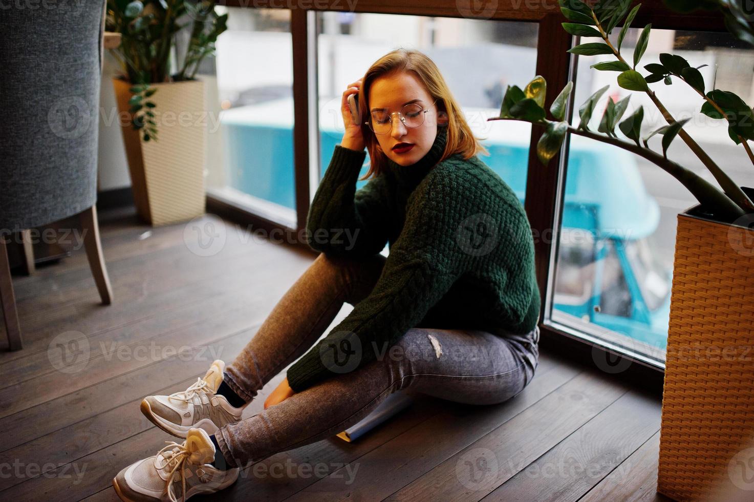 Cheerful young beautiful redhaired woman in glasses, green warm wool sweater, using her notebook, while sitting on cafe against window. She sit on floor and think about something. photo