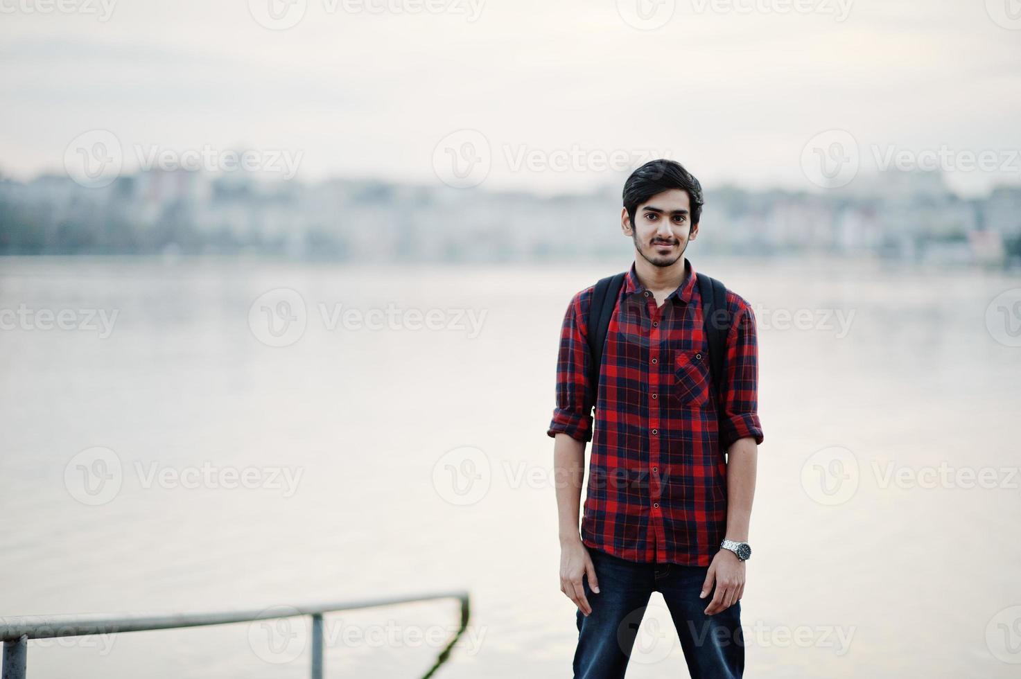 Young indian student man at checkered shirt and jeans with backpack posed on evening city against lake. photo