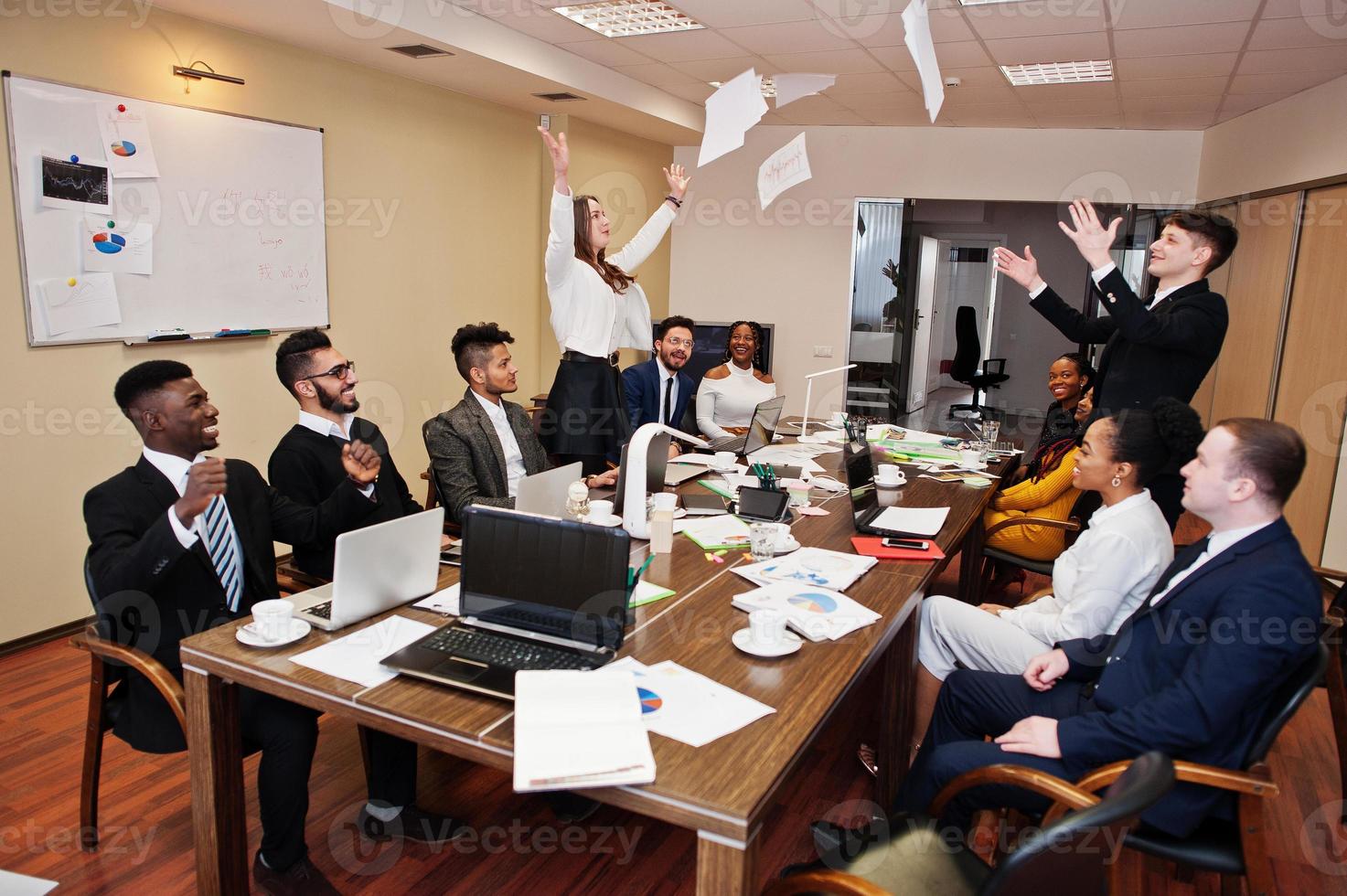 Multiracial business team meeting around boardroom table, two team leaders throw paper up. photo