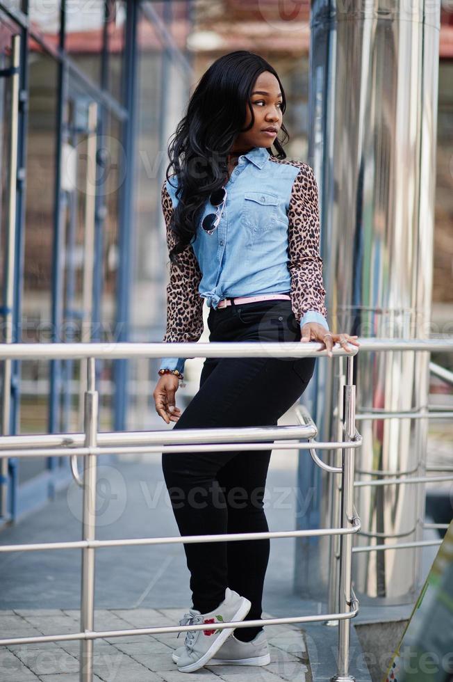 chica afroamericana hipster con camisa de jeans con mangas de leopardo posando en la calle contra un edificio de oficinas moderno con ventanas azules. foto