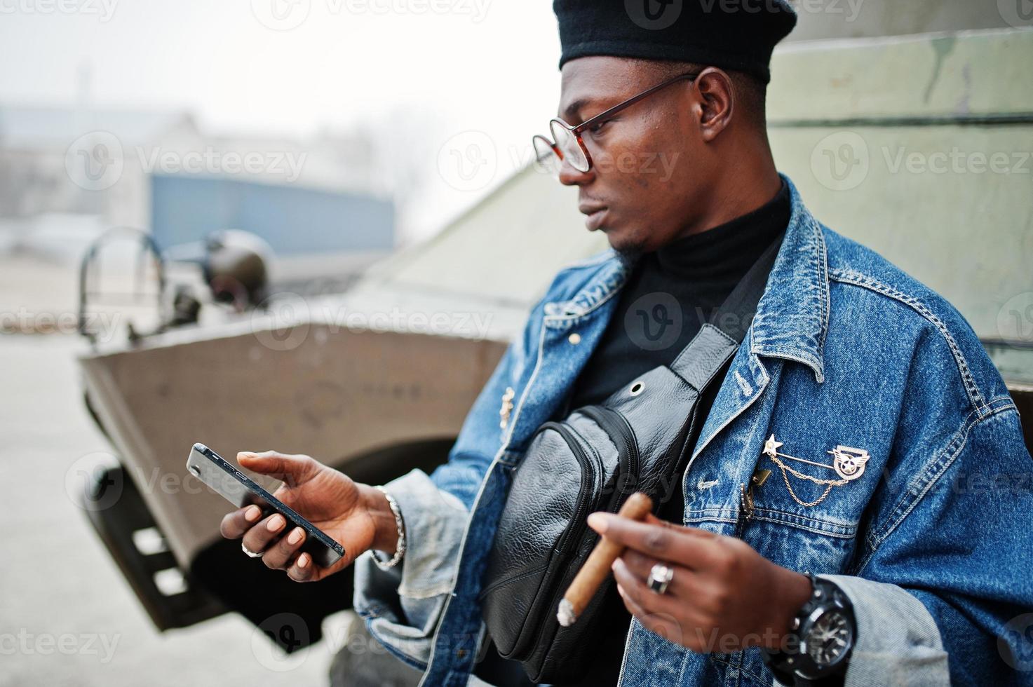 African american man in jeans jacket, beret and eyeglasses, smoking cigar and posed against btr military armored vehicle, with mobile phone at hand. photo