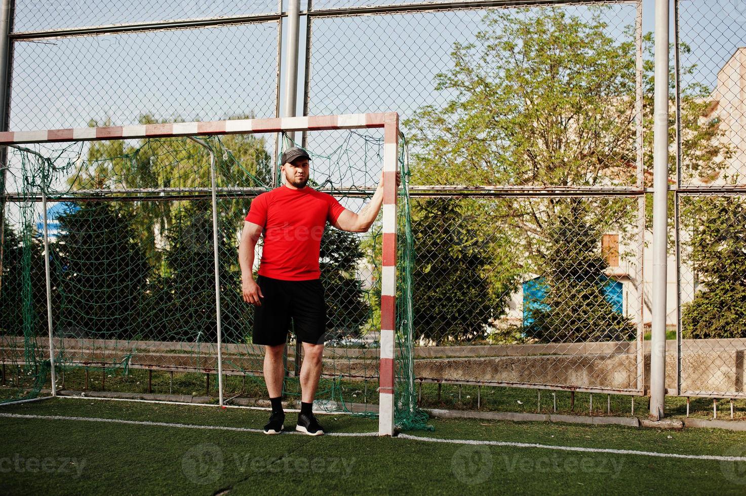 joven musculoso barbudo brutal vestido con camisa roja, pantalones cortos y gorra en el estadio. foto