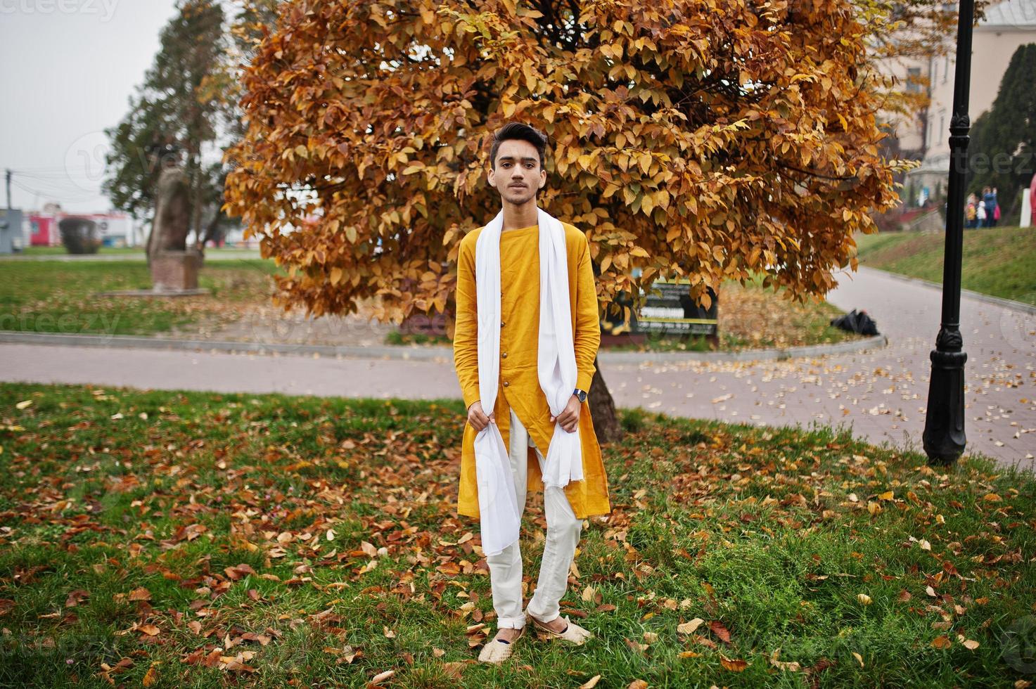 hombre indio con estilo en ropa tradicional amarilla con bufanda blanca posada al aire libre contra las hojas de otoño. foto