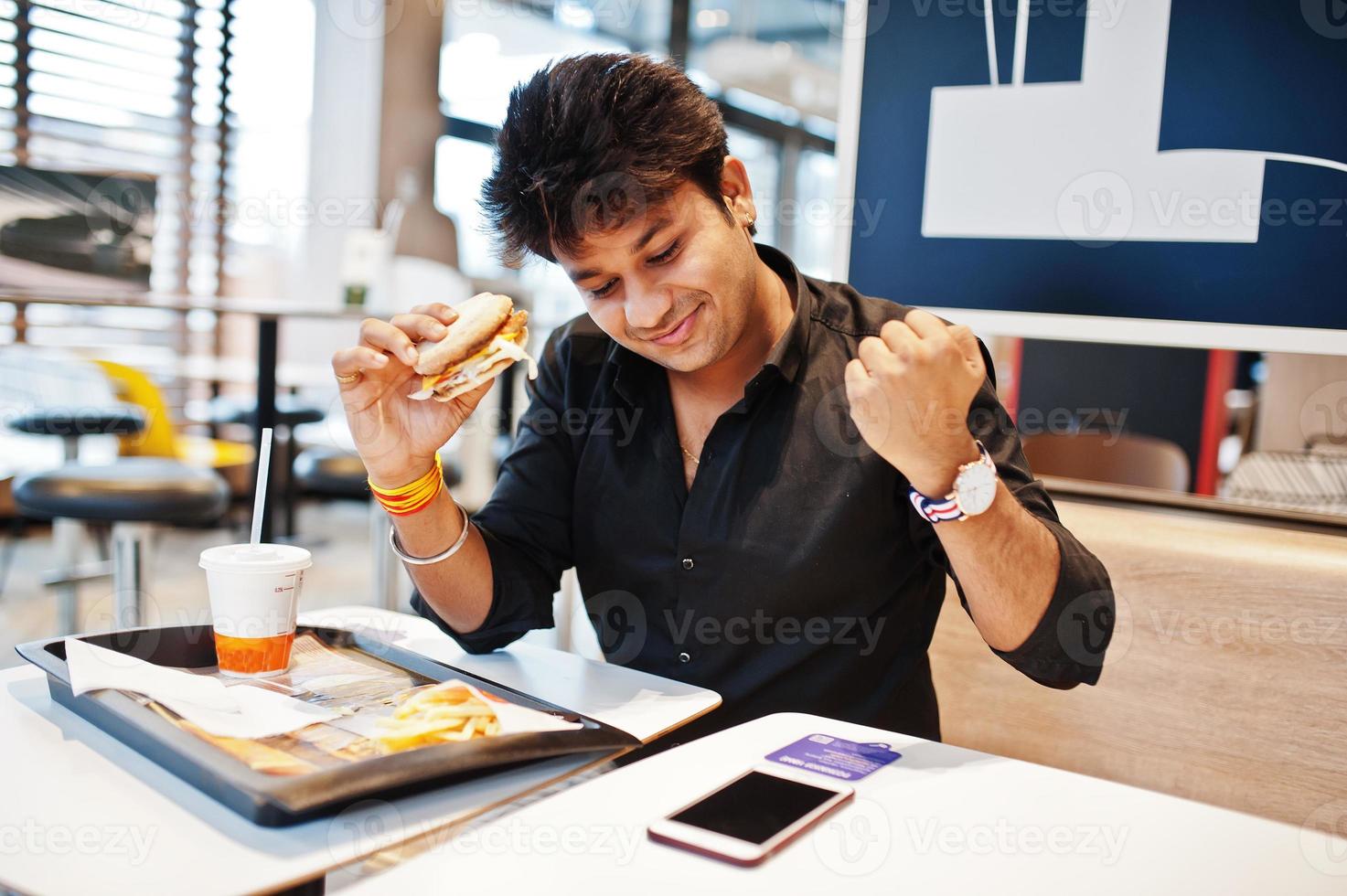 Stylish indian man sitting at fast food cafe and eating hamburger, read morning news on mobile phone. He is happy and show like him win something. photo