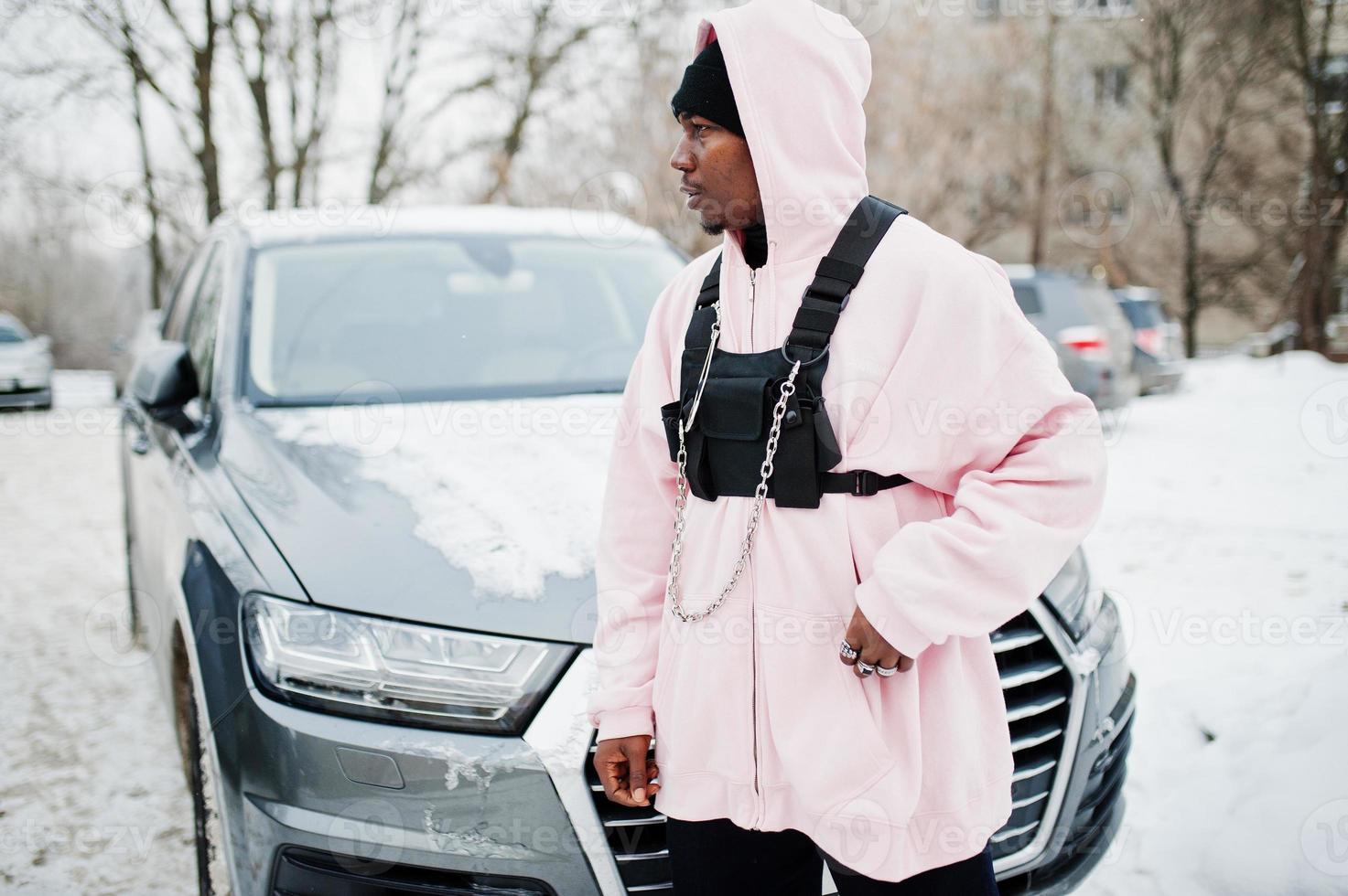 Stylish urban style african american man in pink hoodie posed against suv car at winter. Afro rapper guy. photo