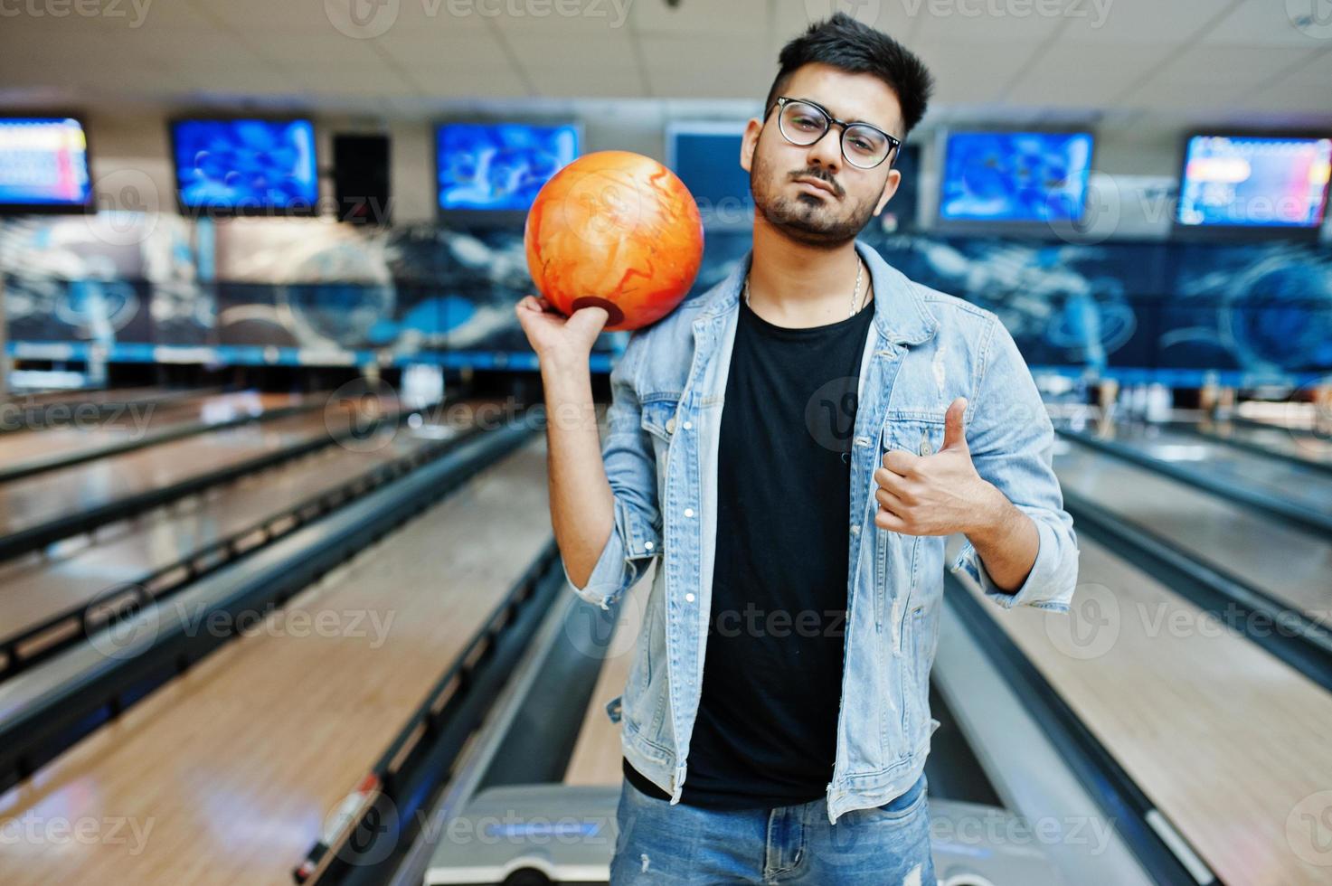 hombre asiático de barba elegante con chaqueta de jeans y gafas de pie en la bolera con la pelota en la mano. foto