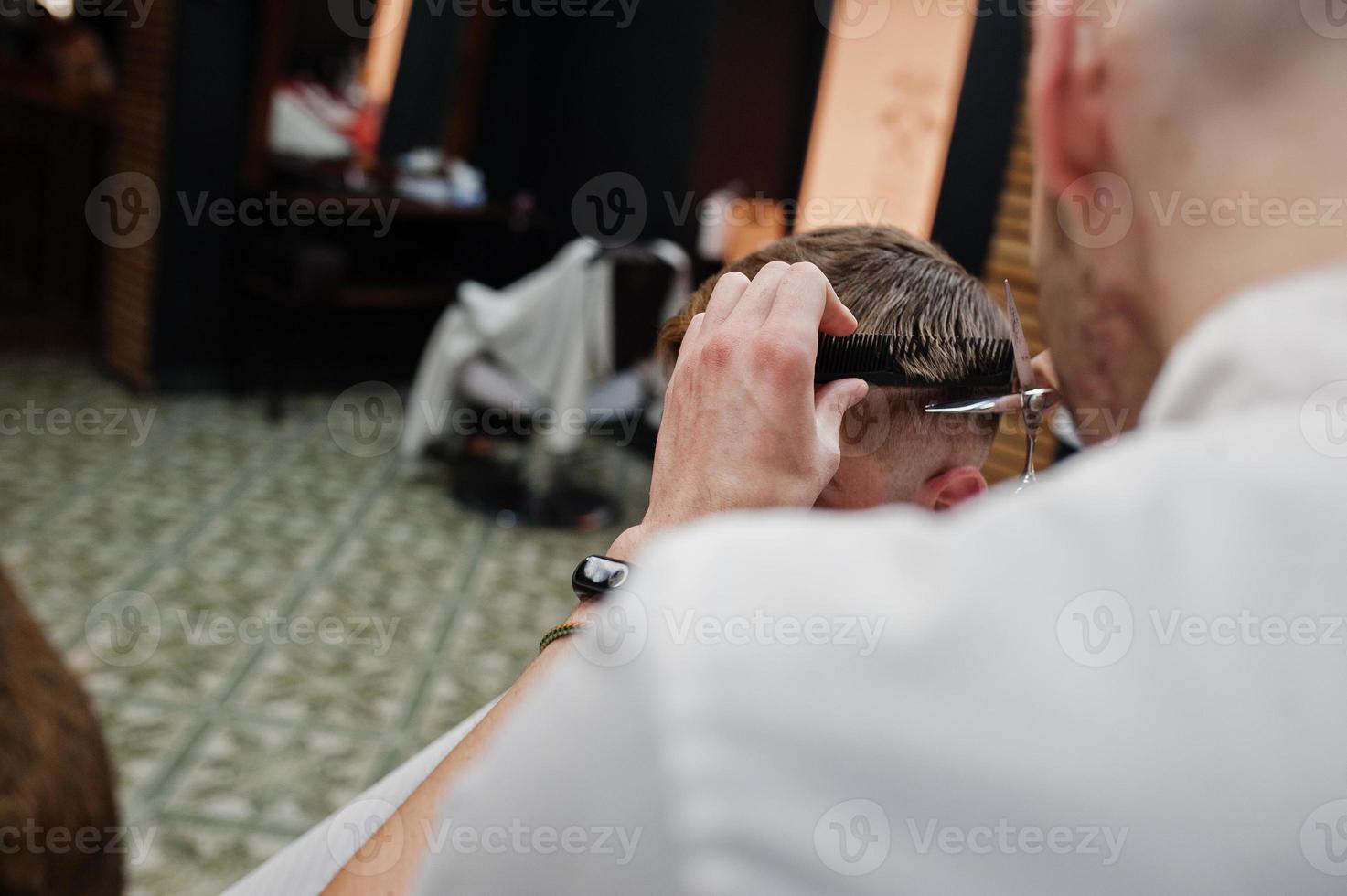 Young bearded man getting haircut by hairdresser while sitting in chair at barbershop. Barber soul. photo