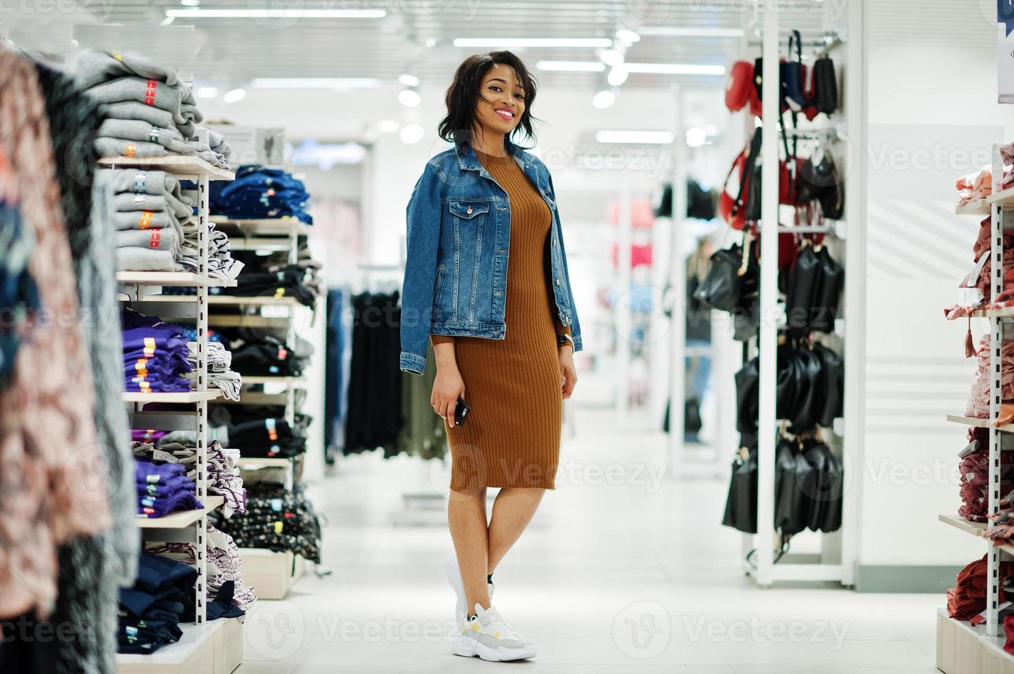 Chic african american woman in brown tunic dress and jeans jacket posed at clothes store. It's time for shopping. photo