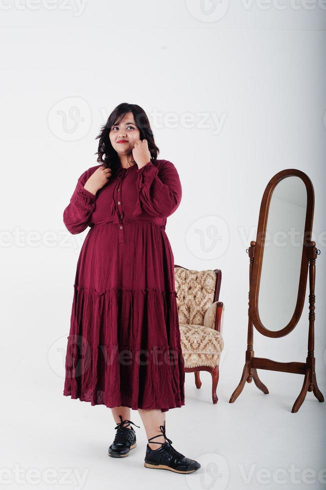 atractiva mujer del sur de asia con un vestido rojo profundo posó en el estudio sobre fondo blanco contra el espejo y la silla. foto