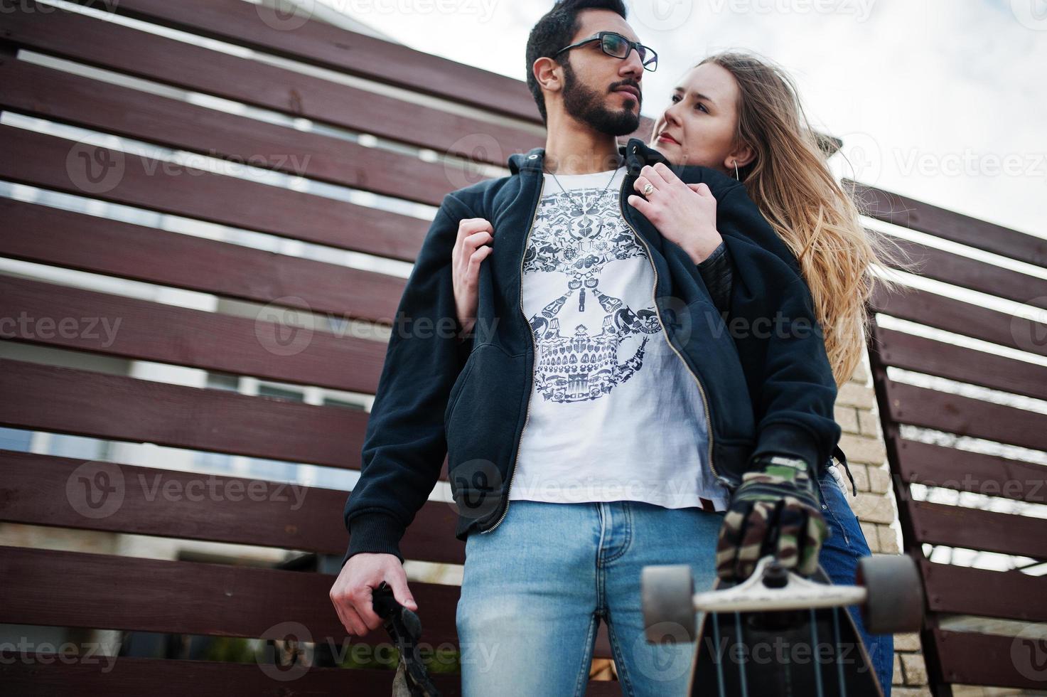 Cool multiracial couple hugs together with longboard against wooden fence. photo