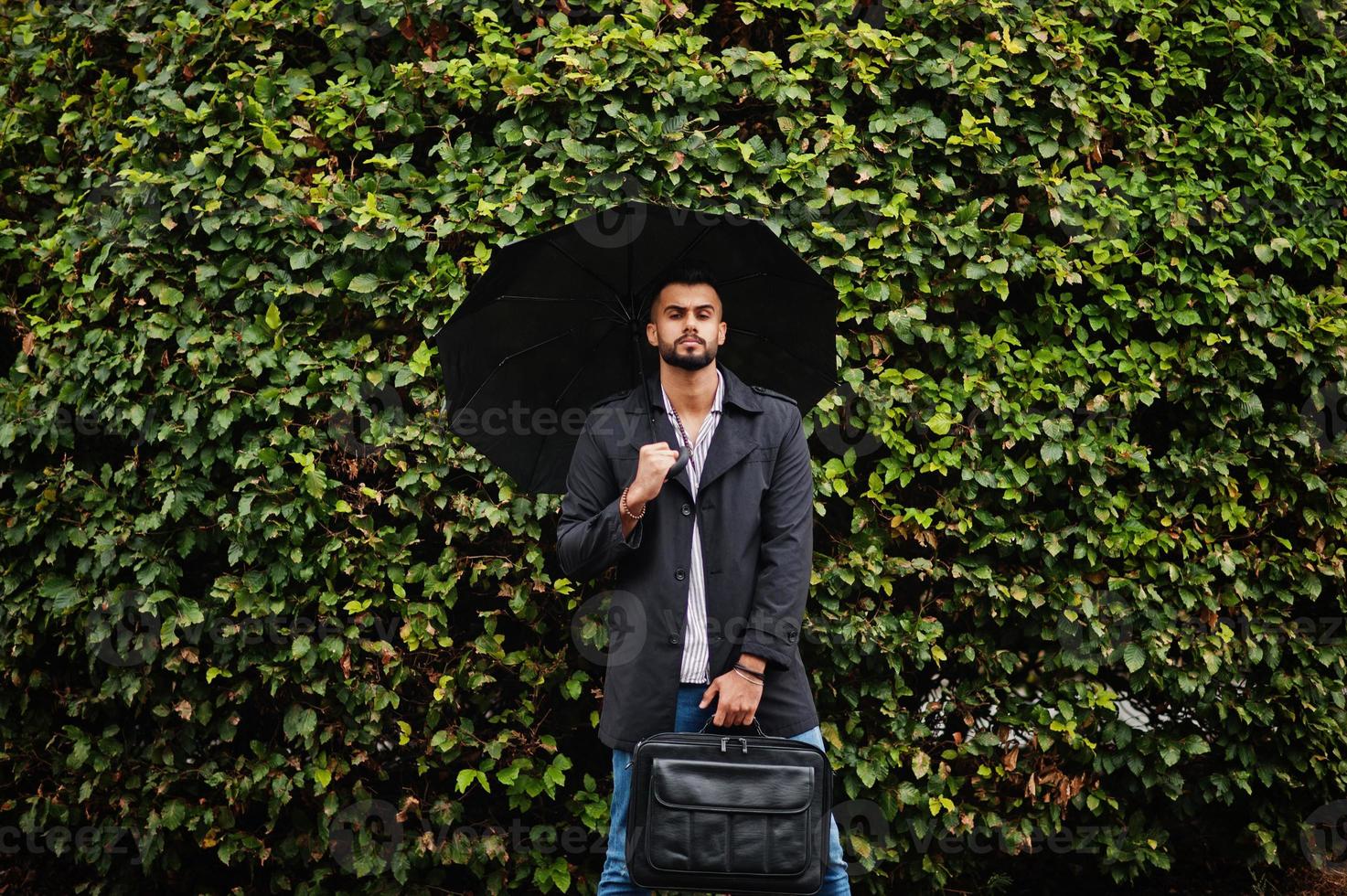 Fashionable tall arab beard man wear on black coat with umbrella and bag case posed at rain weather day. photo