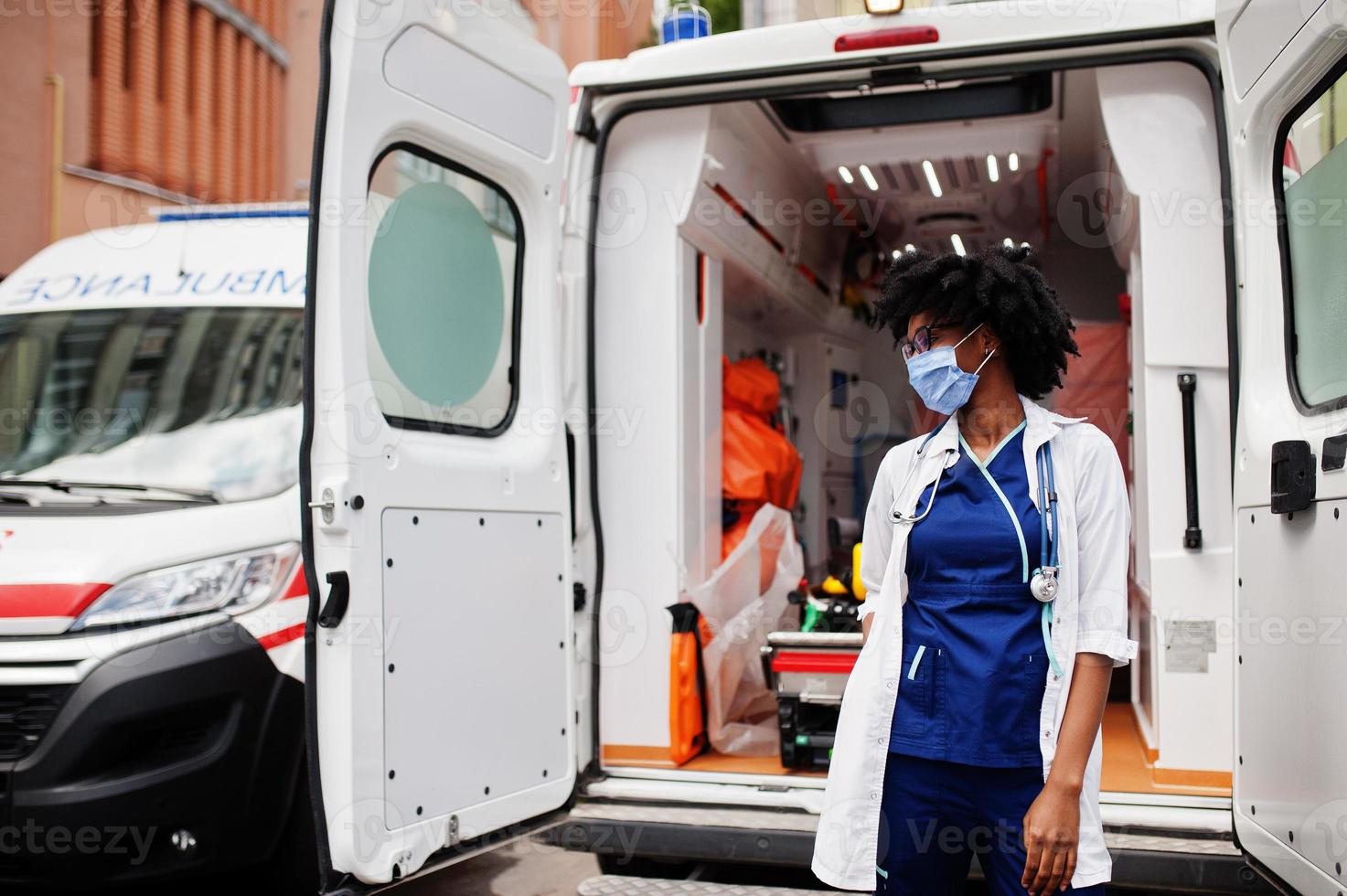 African american female paramedic in face protective medical mask standing in front of ambulance car. photo