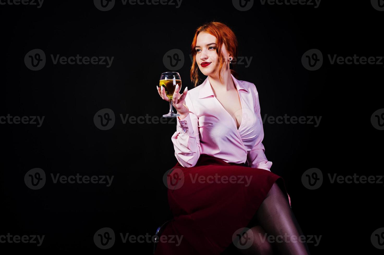 Gorgeous red haired girl in pink blouse and red skirt with glass of wine at hand isolated on black. photo