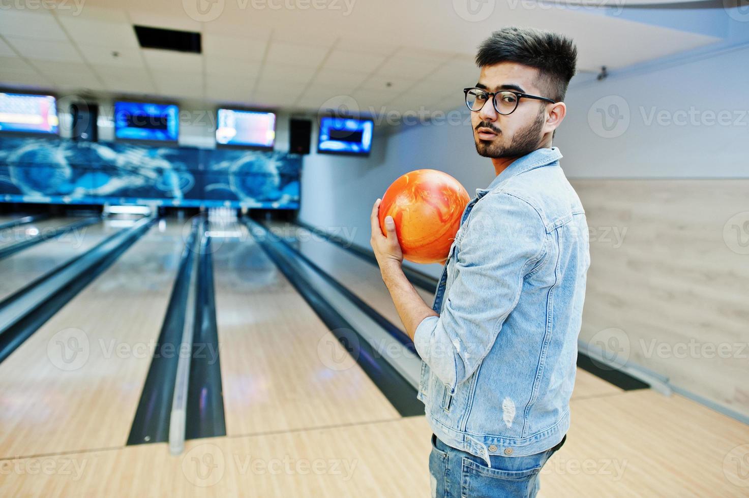 hombre asiático con estilo en chaqueta de jeans y gafas de pie en la bolera con la pelota en la mano. foto