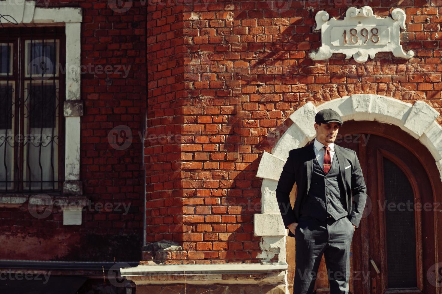 Portrait of retro 1920s english arabian business man wearing dark suit, tie and flat cap standing against old brick house 1898 year. photo